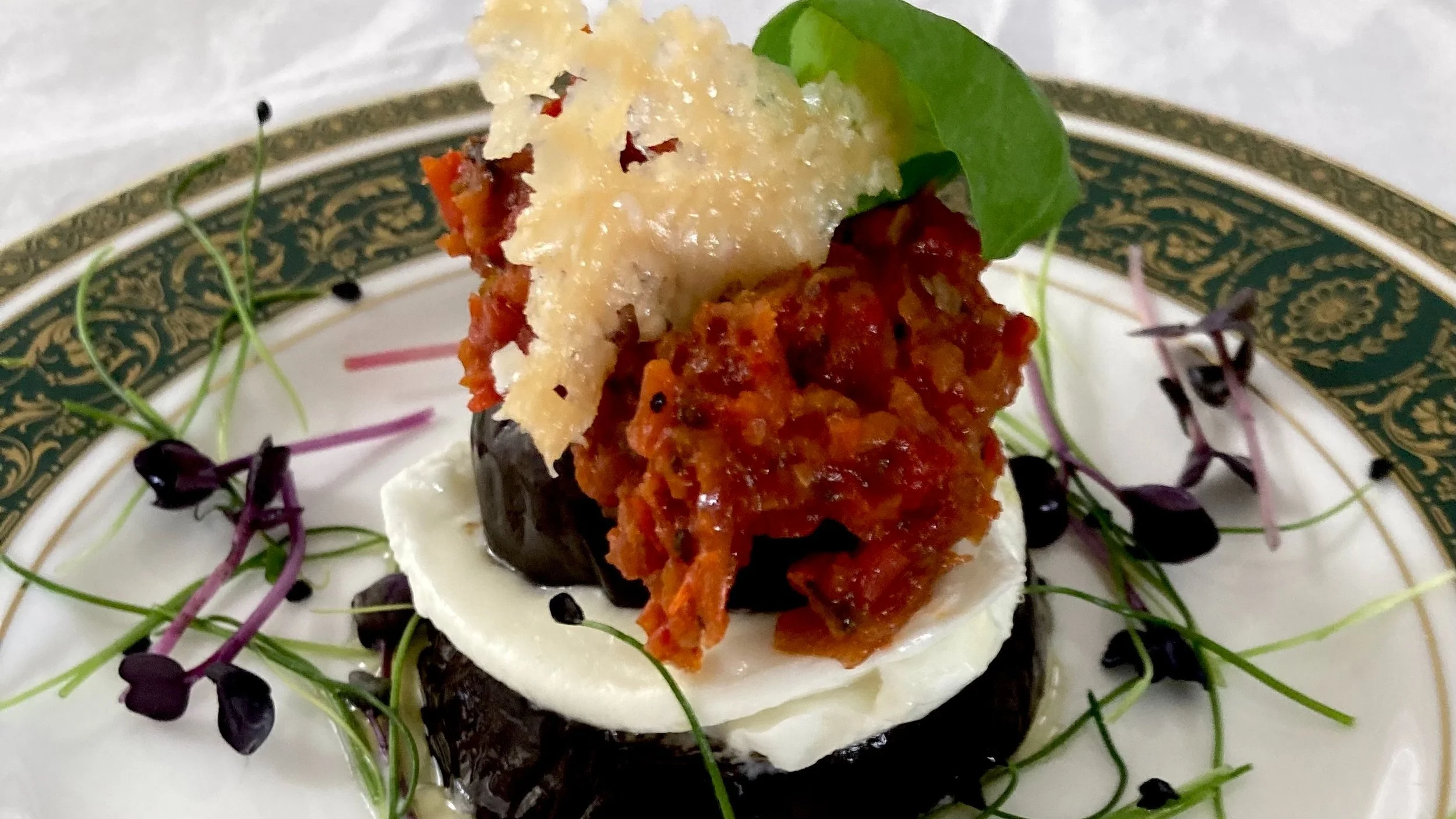 A gourmet appetizer featuring a slice of roasted eggplant topped with a dollop of red sauce, a piece of mozzarella cheese, and a spoonful of cooked minced vegetables topped with shaved cheese, garnished with microgreens on a decorative plate.