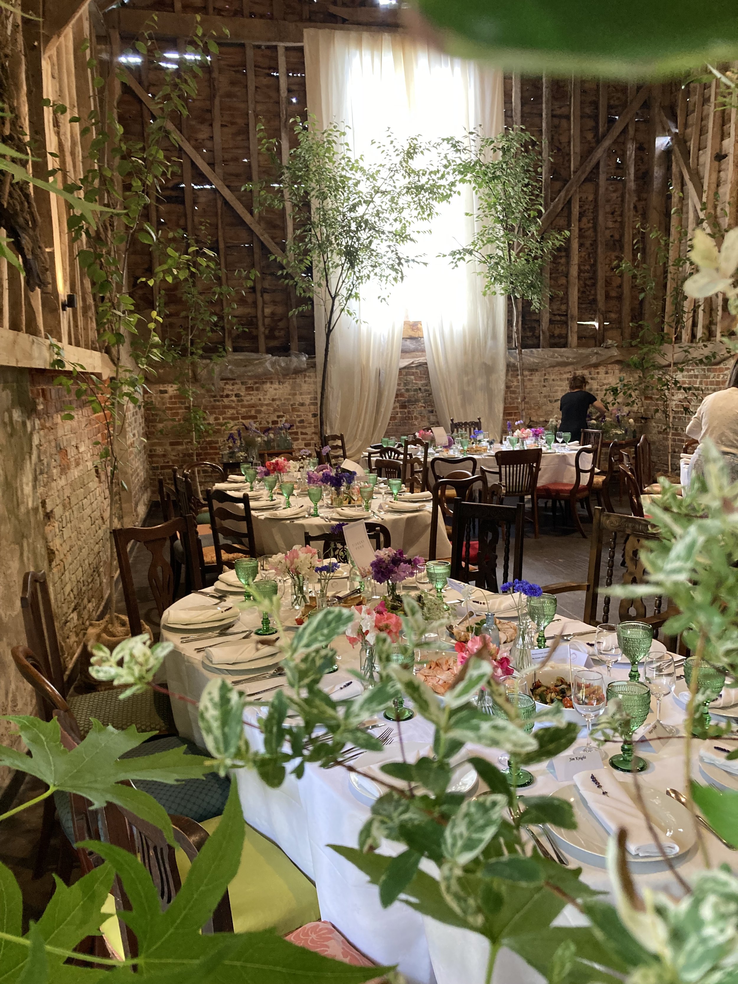 A rustic indoor event space with decorated tables set for a celebration, featuring floral centerpieces, glassware, and tableware, with natural light coming through a large window covered with white curtains, exposed brick walls, and wooden beams.