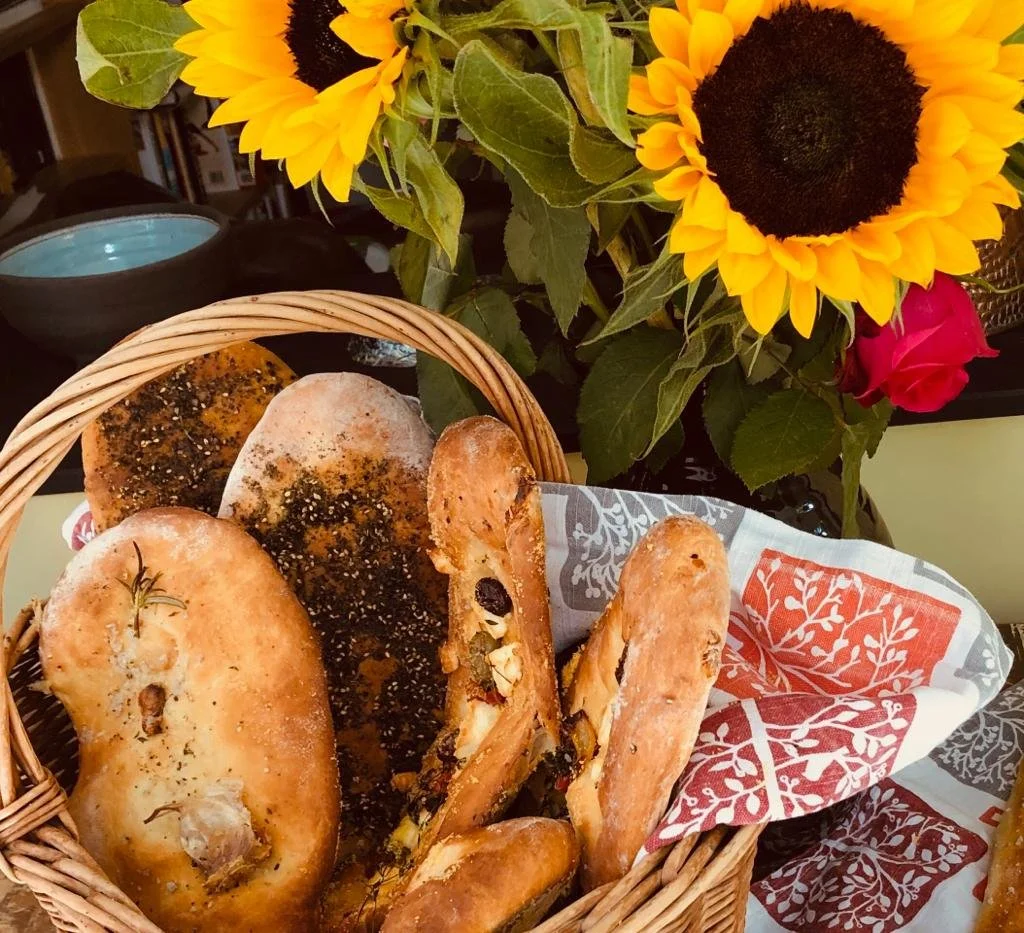 A basket of baked bread with different shapes and toppings, placed on a table with a floral napkin. In the background, a bouquet of vibrant sunflowers and a pink rose.