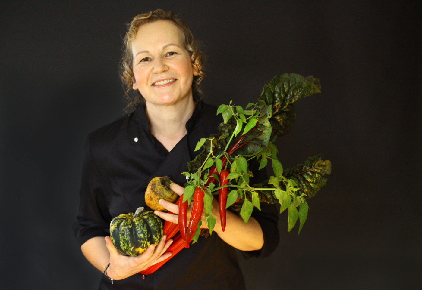 Smiling woman in black attire holding a bouquet of fresh vegetables including a pumpkin, tomatoes, and peppers against a dark background.