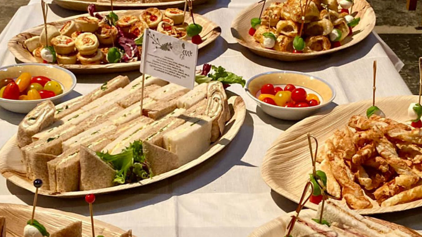 A spread of finger sandwiches, cherry tomatoes, bruschetta, mini pizzas, and meats on wooden served trays and plates, with some garnished with pickles, small greens, and toothpicks.