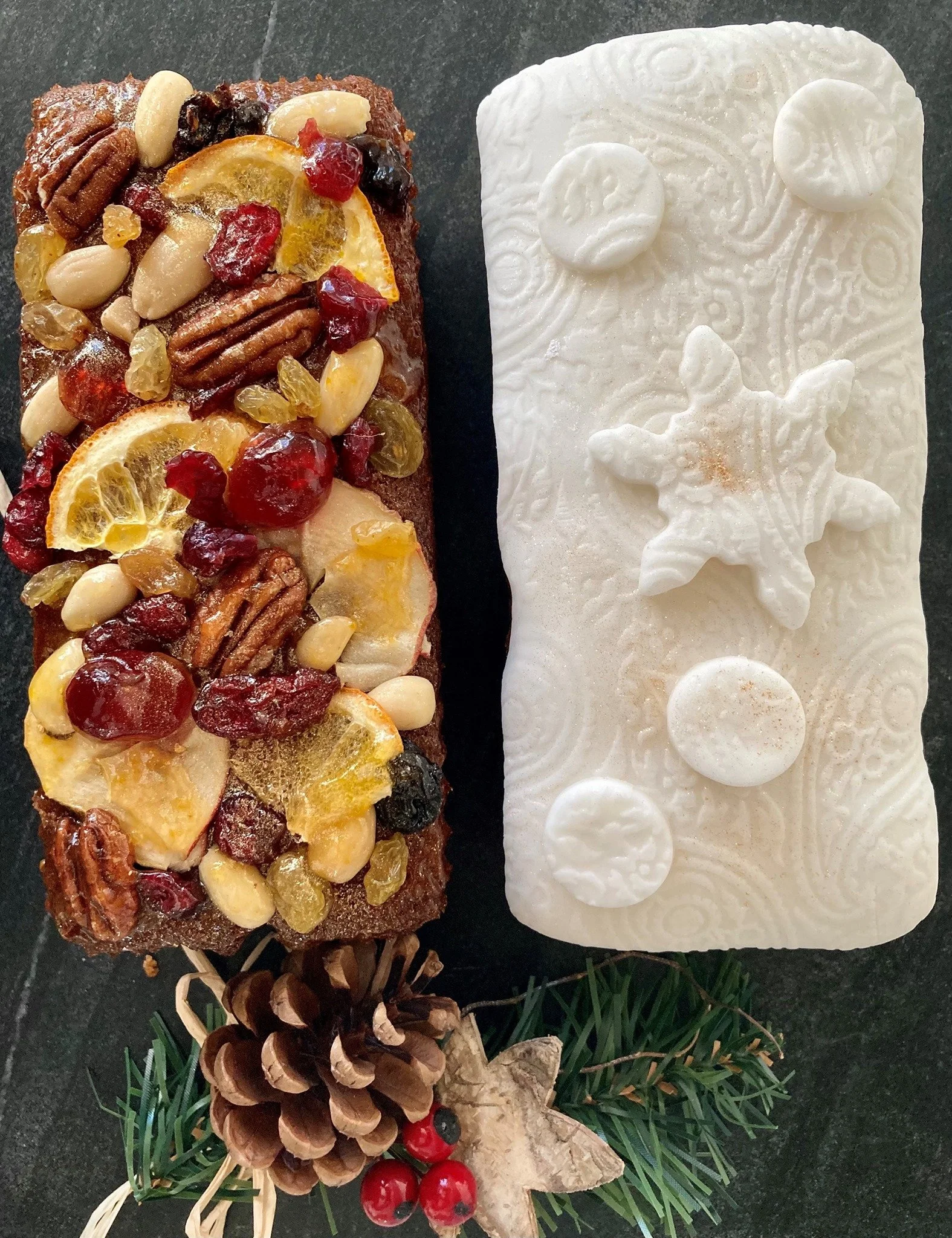 Christmas-themed dessert resembling a log, with candied fruit, nuts, and edible decorations, next to a white decorative snowflake-shaped pattern with raised circles and swirls, on a dark surface, decorated with pinecones, holly berries, and evergreen