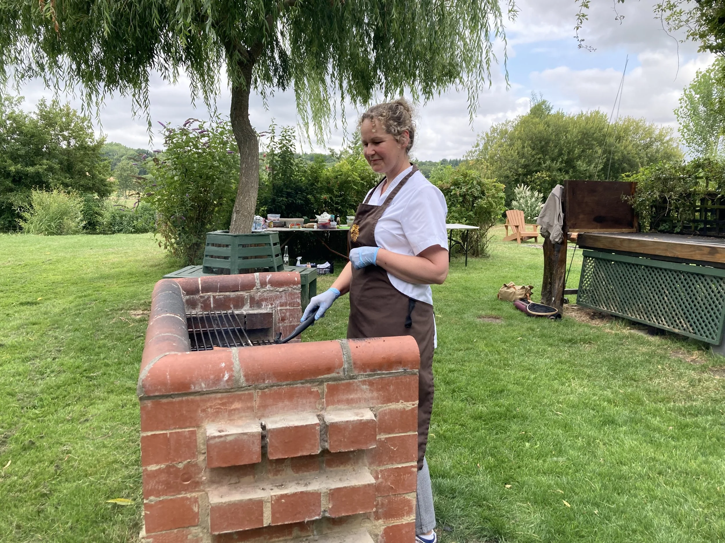 A woman grilling outdoors on a brick barbecue grill, wearing a brown apron, white shirt, and disposable gloves, with green grass, trees, and outdoor furniture in the background.