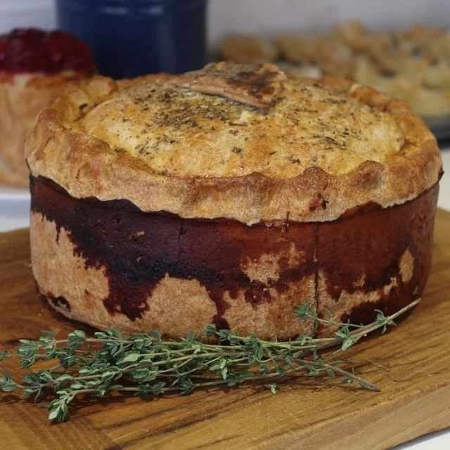 A savory pie with a golden-brown crust on a wooden cutting board, garnished with thyme, with a side of jam or fruit spread in the background.