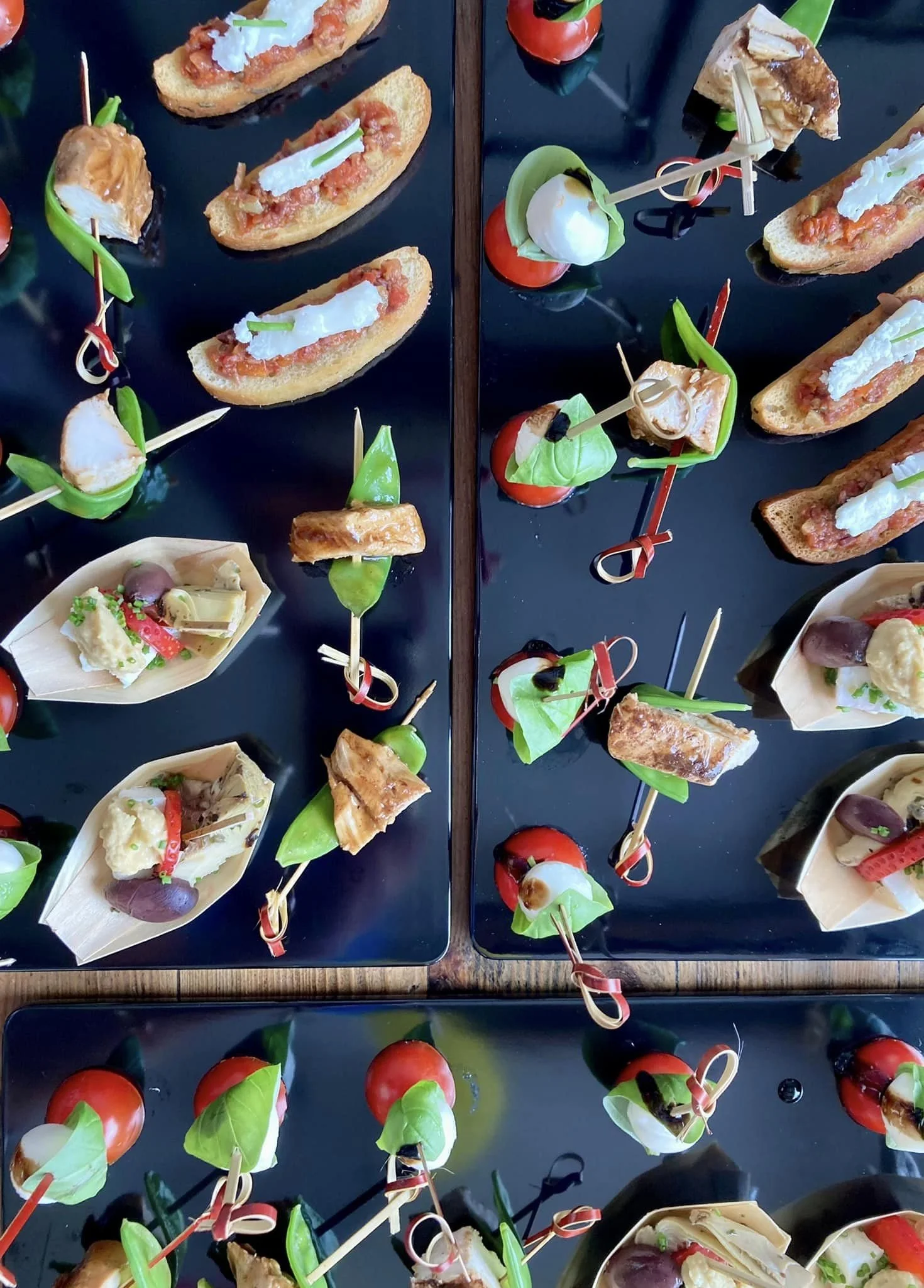 Assorted bite-sized gourmet appetizers arranged on black trays, including bruschetta with tomatoes and cheese, and other small snacks with vegetables, cheese, and cured meats, garnished with fresh herbs and small skewers.