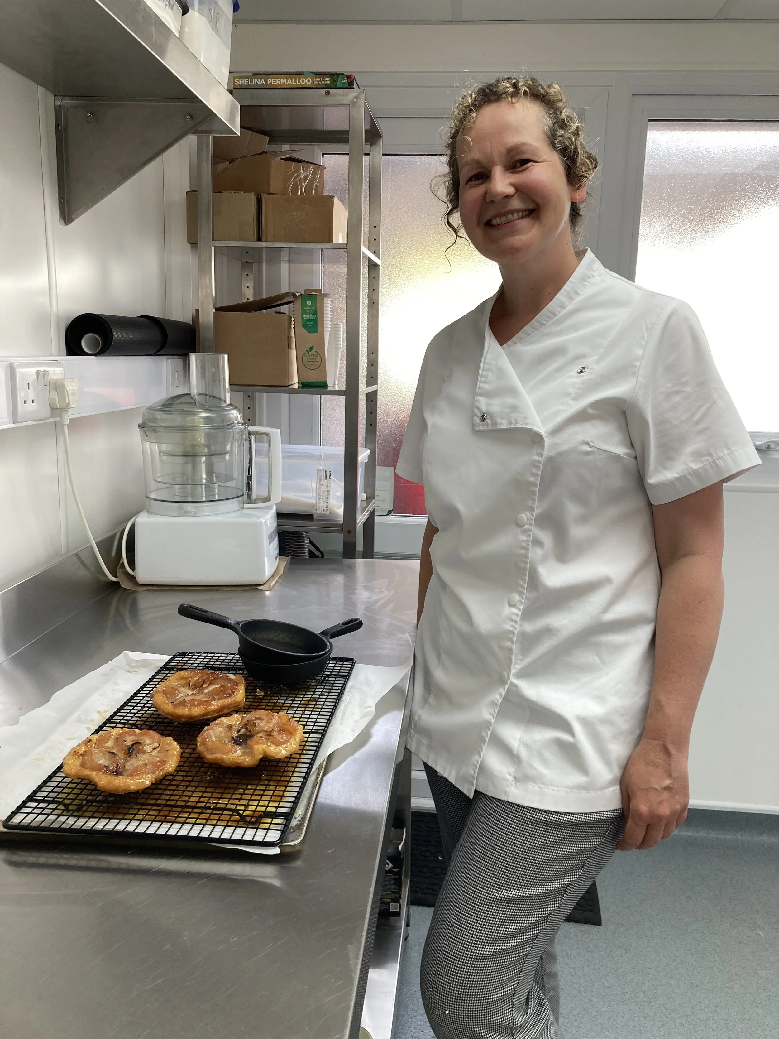 A woman in a white chef's coat standing in a kitchen next to a cooling rack with three pastries on it, smiling at the camera.