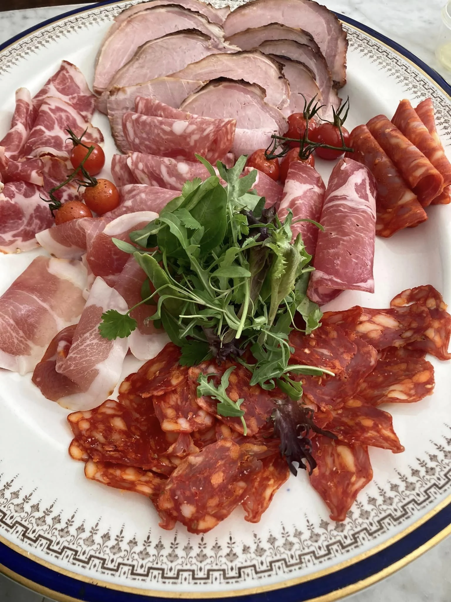 An assortment of cured meats, cherry tomatoes, and mixed greens on a decorative white plate.
