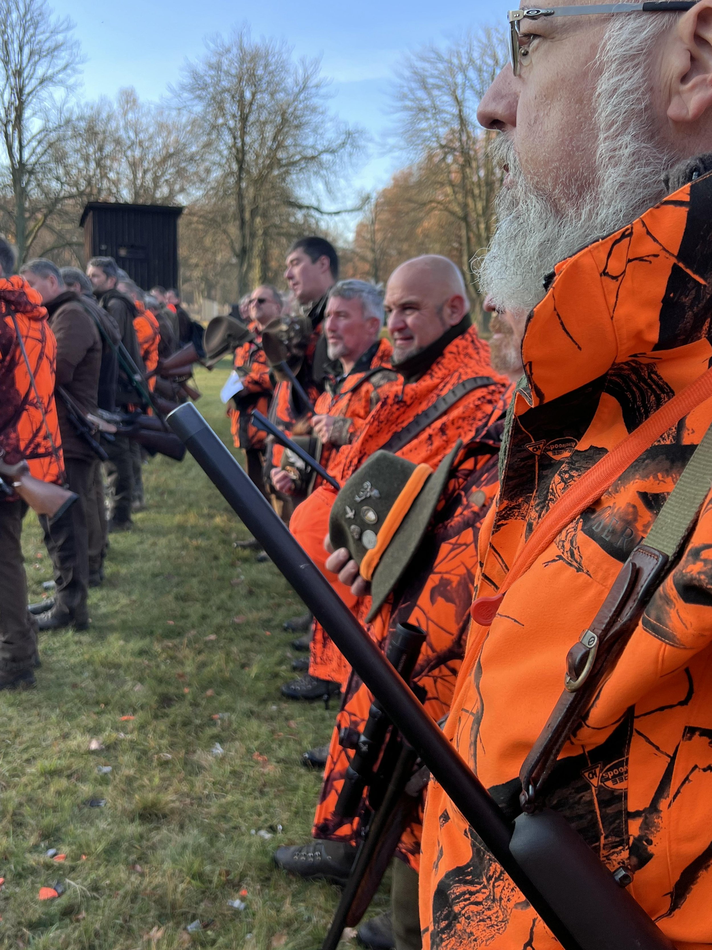 Gruppo di persone con abbigliamento da caccia, alcune con fucili, accompagnate da un cane, in un ambiente naturale.