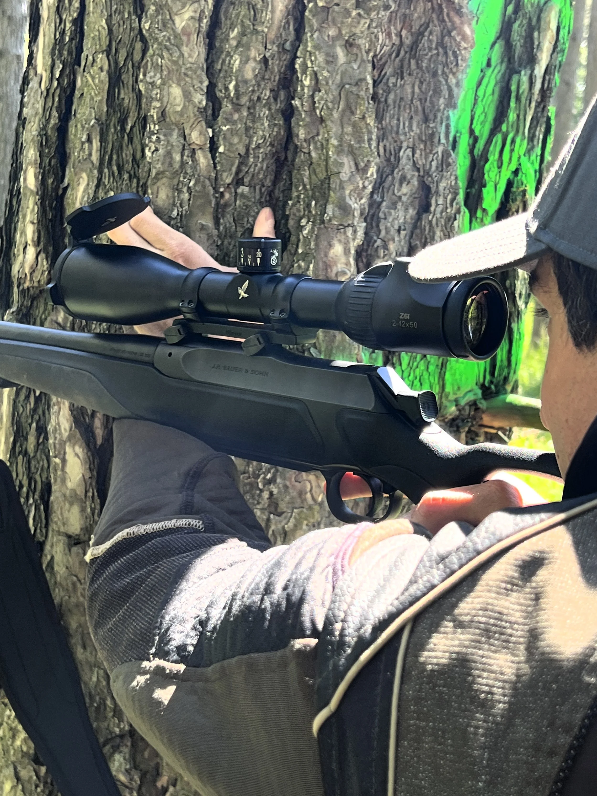 Uomo con cappello da cacciatore che usa un fucile con cannocchiale in una foresta, vicino a un albero.
