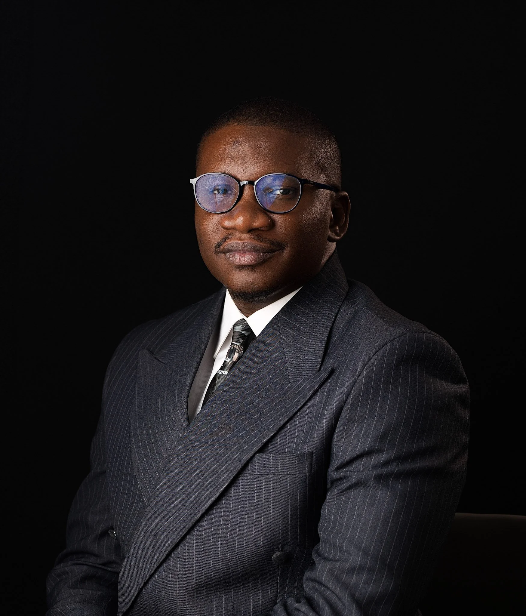 A man wearing glasses and a black suit sits at a wooden table with his hands folded, wearing a white shirt, a wristwatch, and a ring, in a professional setting.