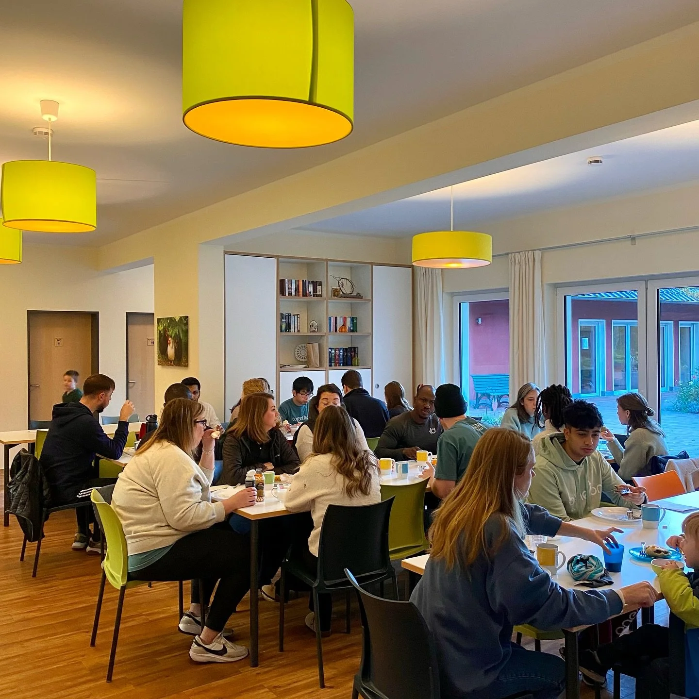 Church members sitting at tables in a bright dining area with green pendant lights and white curtains during our autumn retreat for International Church Berlin.