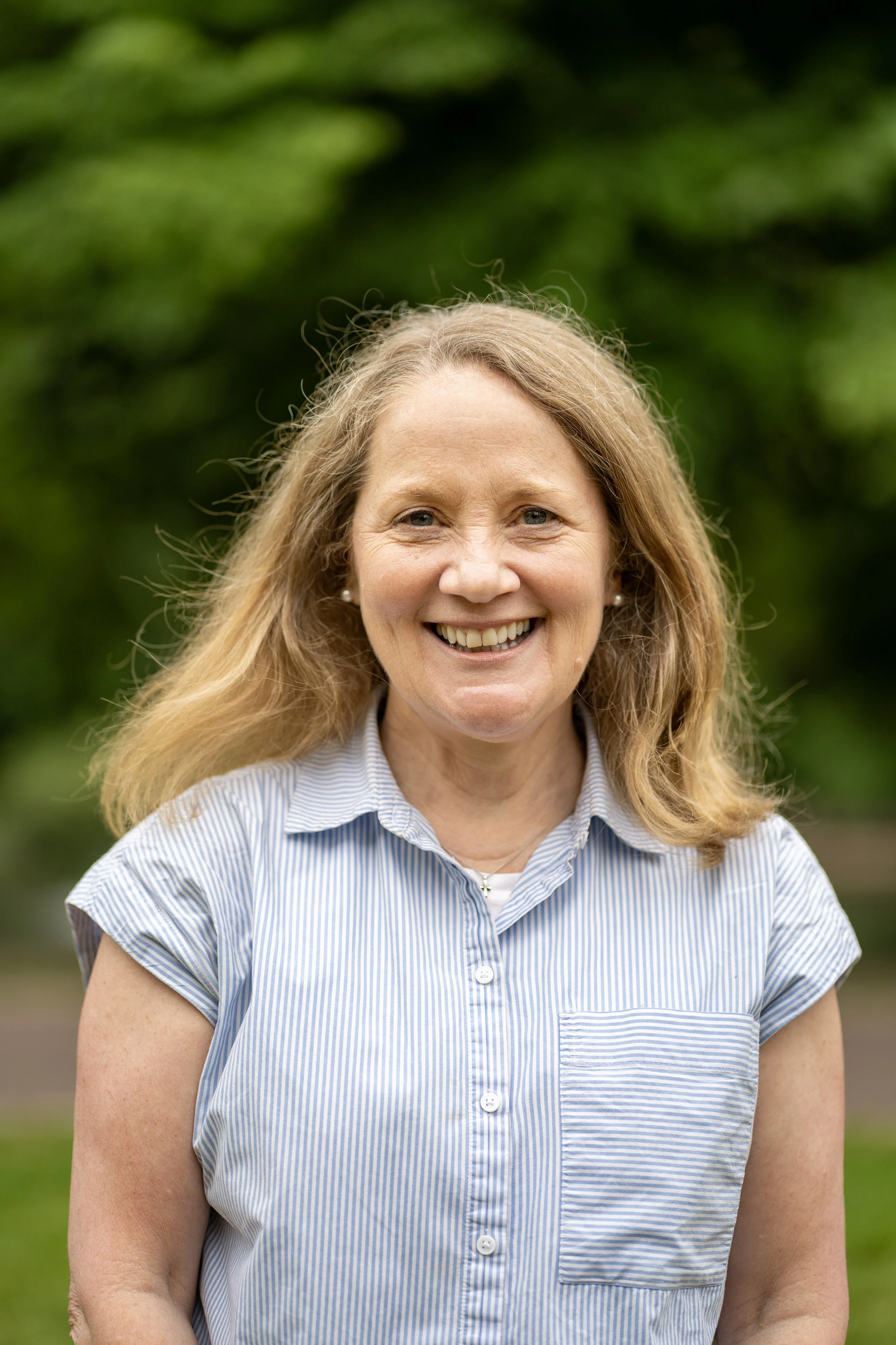 A smiling middle-aged woman with blonde hair wearing a blue and white striped short-sleeve shirt outdoors with green trees in the background.