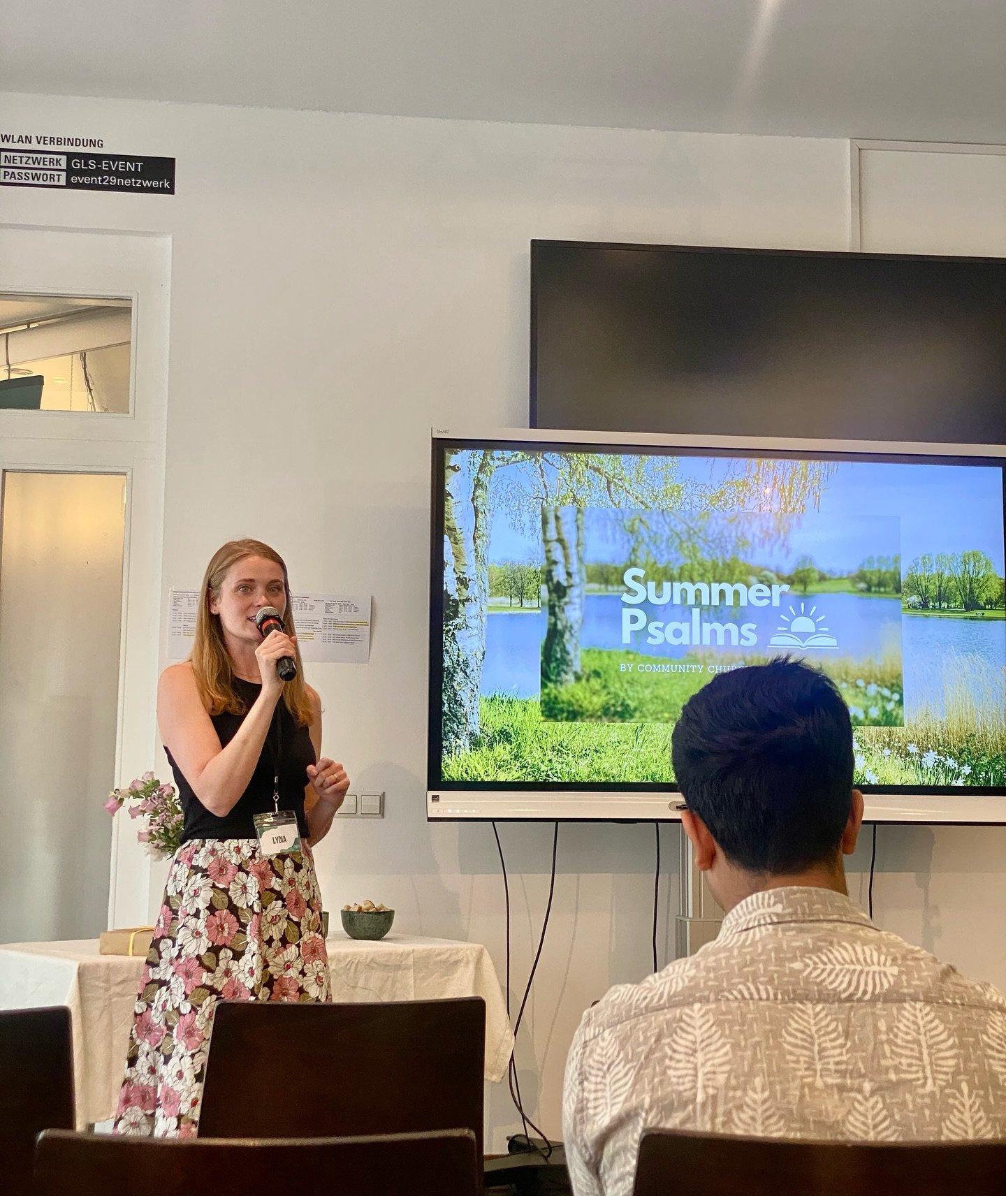 Pastor Lydia Whitley preaching the 'Summer Psalms' series at our English church Berlin, speaking into a microphone with a man seated in front and a large presentation screen behind her.