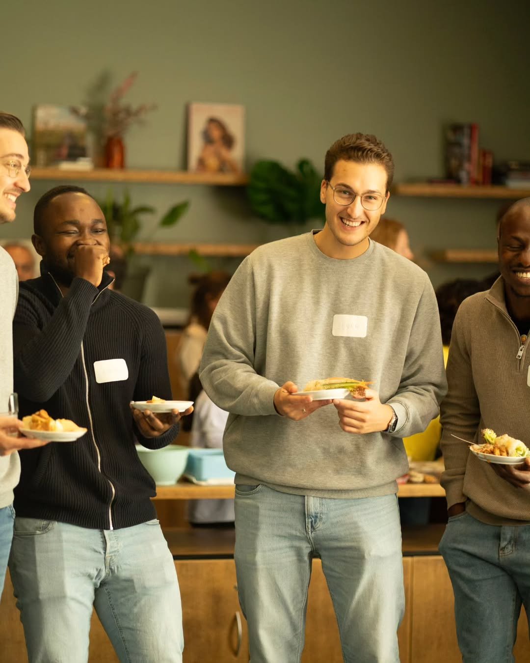 A group of young adults enjoying a social gathering, holding plates of food and smiling in a cozy room with bookshelves and artwork in the background.