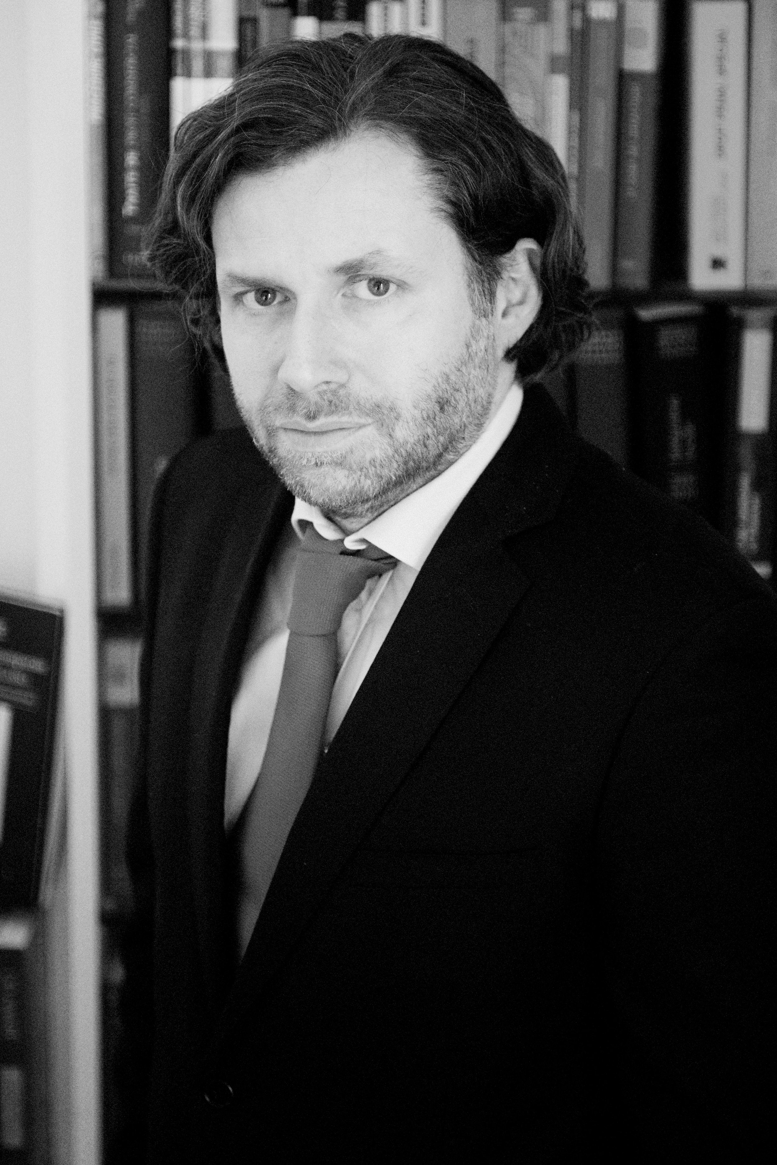 Homme avec des livres en arrière-plan, portant un costume et une cravate.