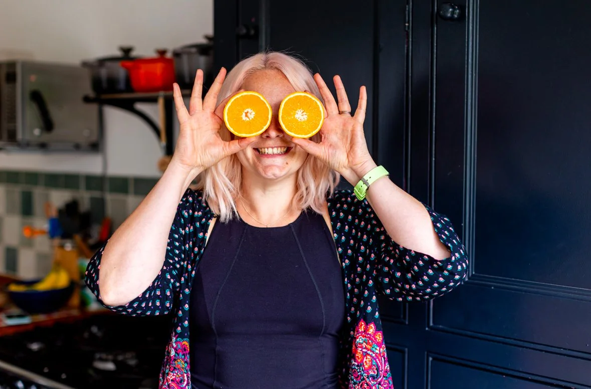 Ruth is stood holding 2 slices of juicy orange over her eyes with a big smile on her fsce