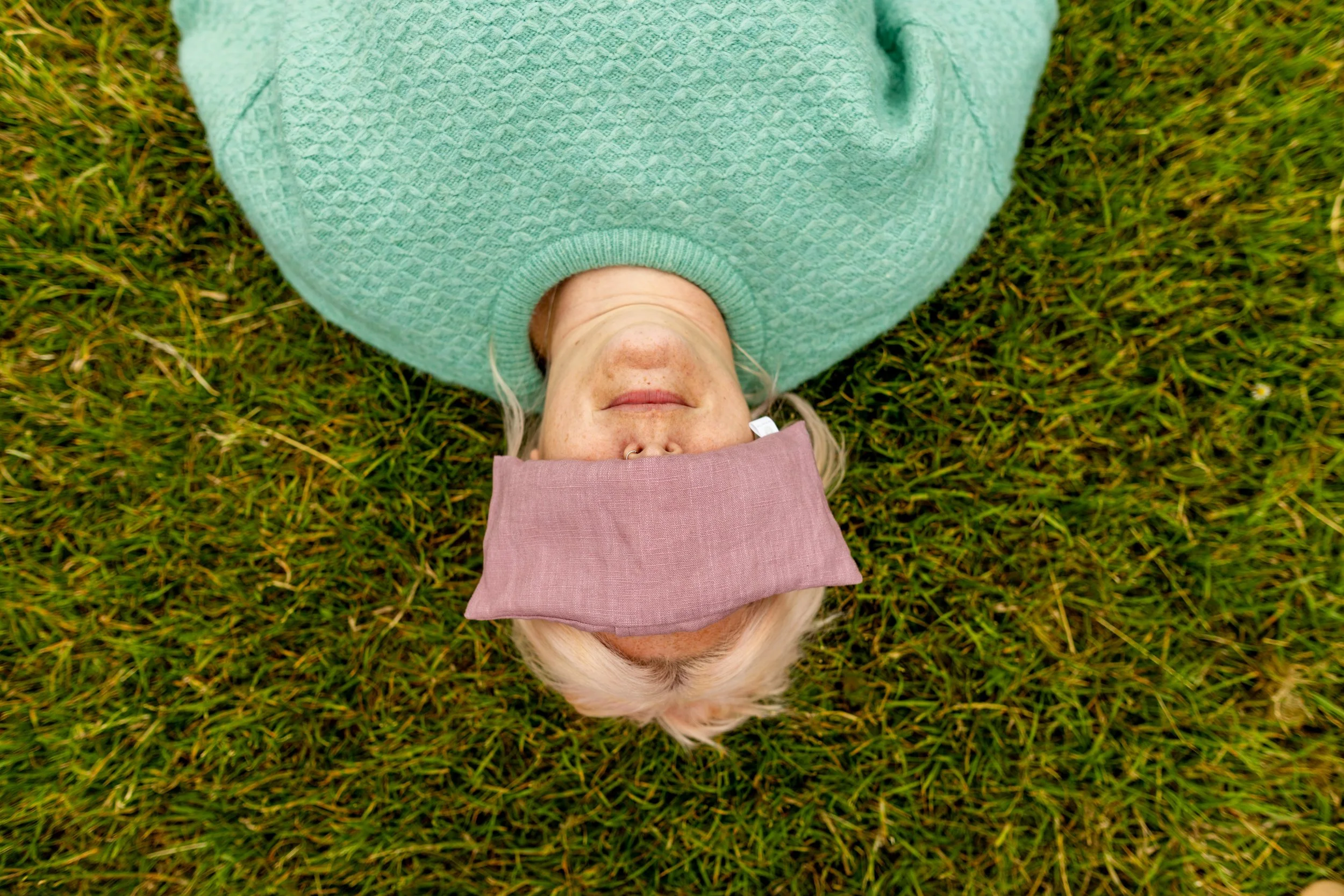 Ruth is relaxing on the grass in Savasana, wearing a mint green jumper and a eye pillow over her eyes