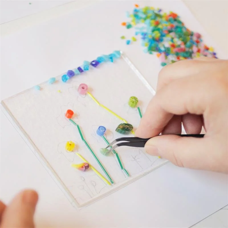 A hand is holding a pair of tweezers and placing small pieces of colour glass on a piece of plain glass. Creating a fused glass piece of art