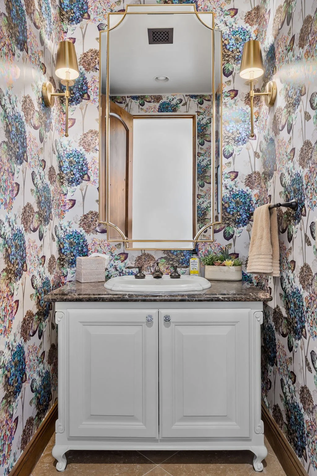 Elegant powder room redesign in Gold Canyon, AZ, featuring hydrangea floral wallpaper, gold sconces, and a classic white vanity by Red Carrot Design.