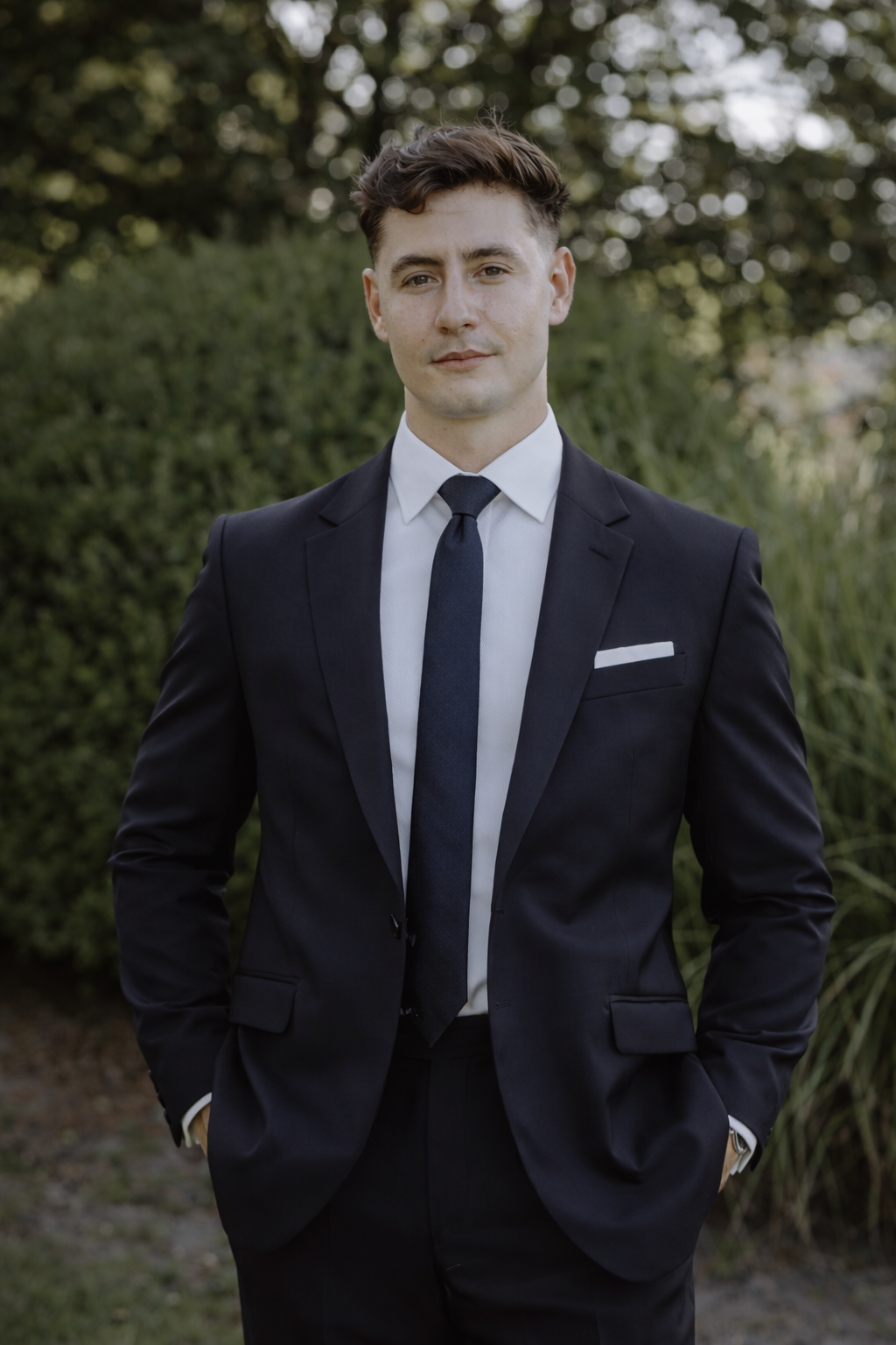 A young man with short brown hair, blue eyes, wearing a dark suit and light blue shirt, smiling outdoors with a blurred background of green trees and cloudy sky.
