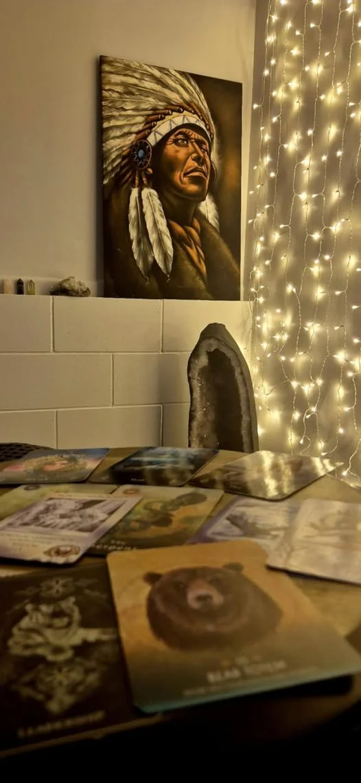 A room with a painting of a Native American chief with a feather headdress on the wall, string lights hanging beside it, a table with various tarot or oracle cards, and a stone or mineral display on a shelf.