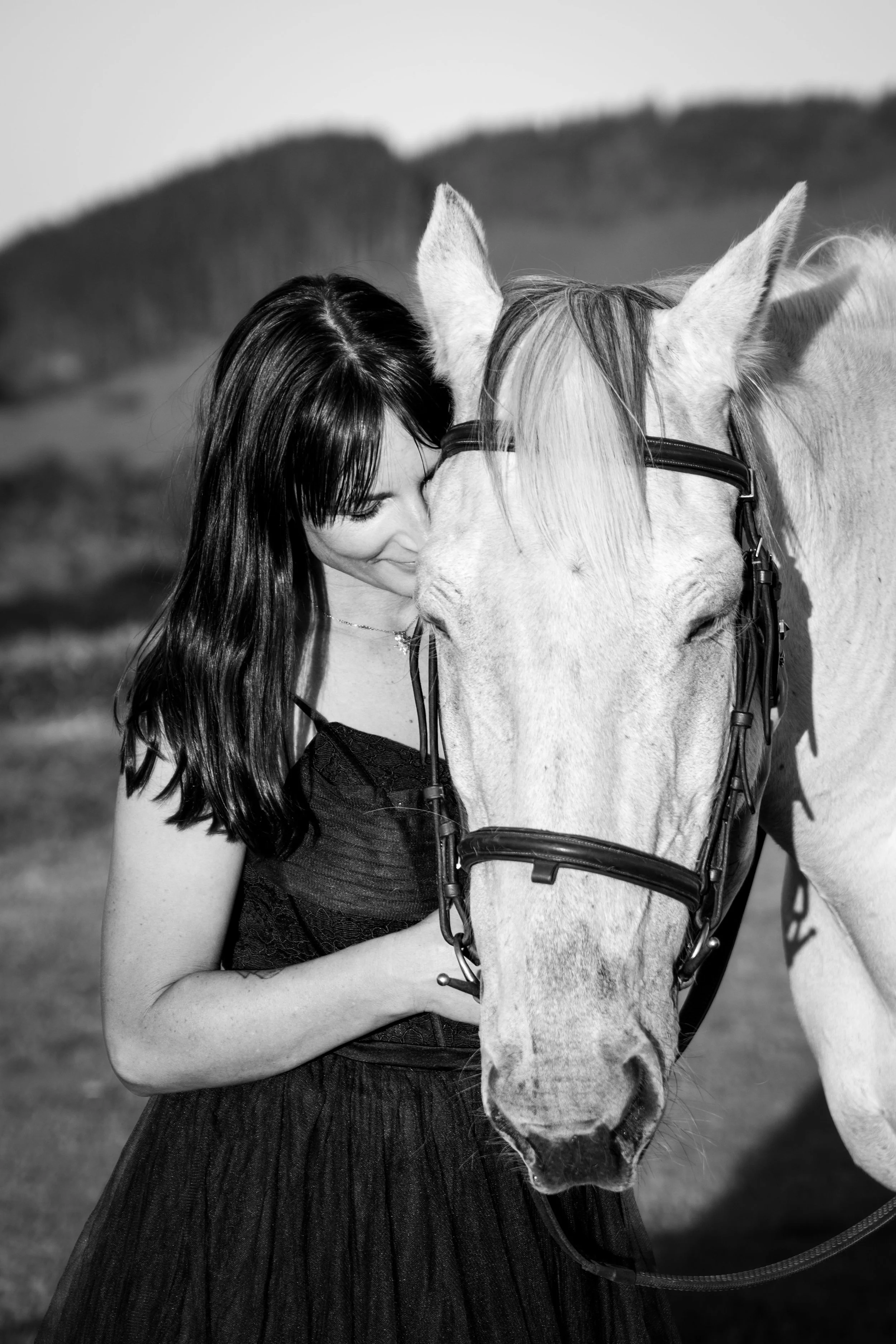 A woman with long dark hair hugging a white horse in an outdoor setting with hills in the background.