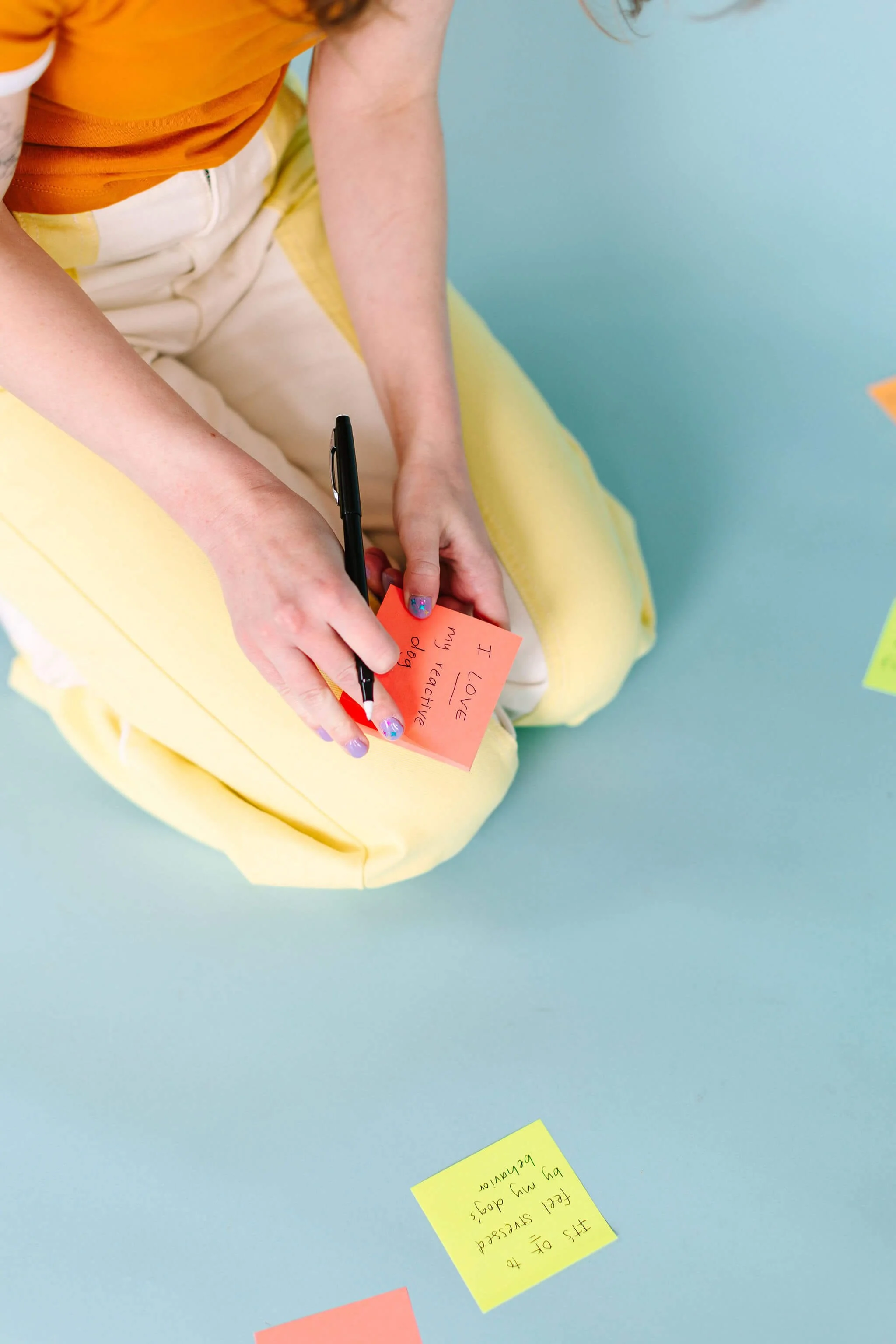 A photo of the lower half of Logan - she kneels on the ground, writing on a sticky note pad propped on her knee.