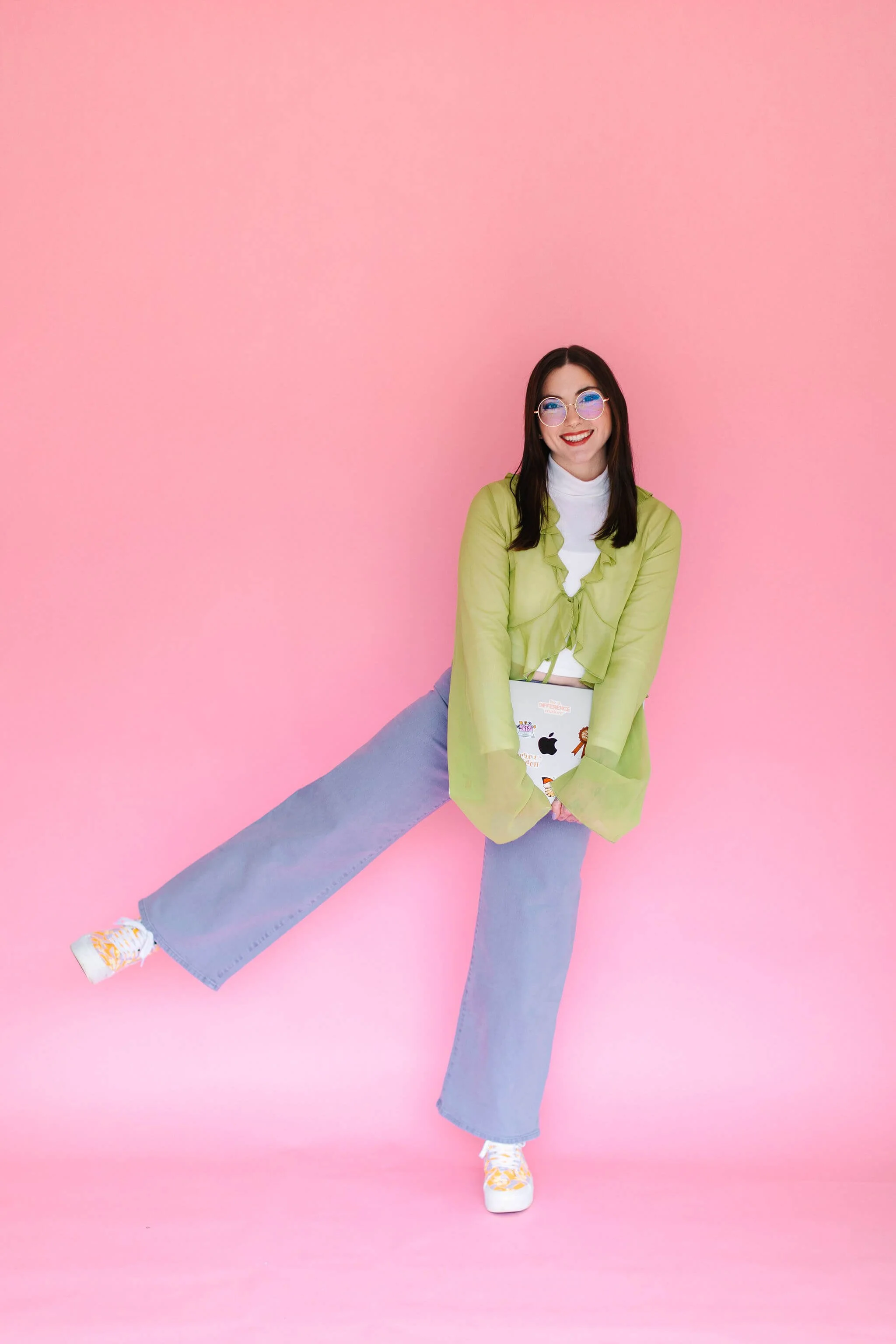 Emily stands in front of a light pink backdrop, holding her laptop and smiling at the camera.