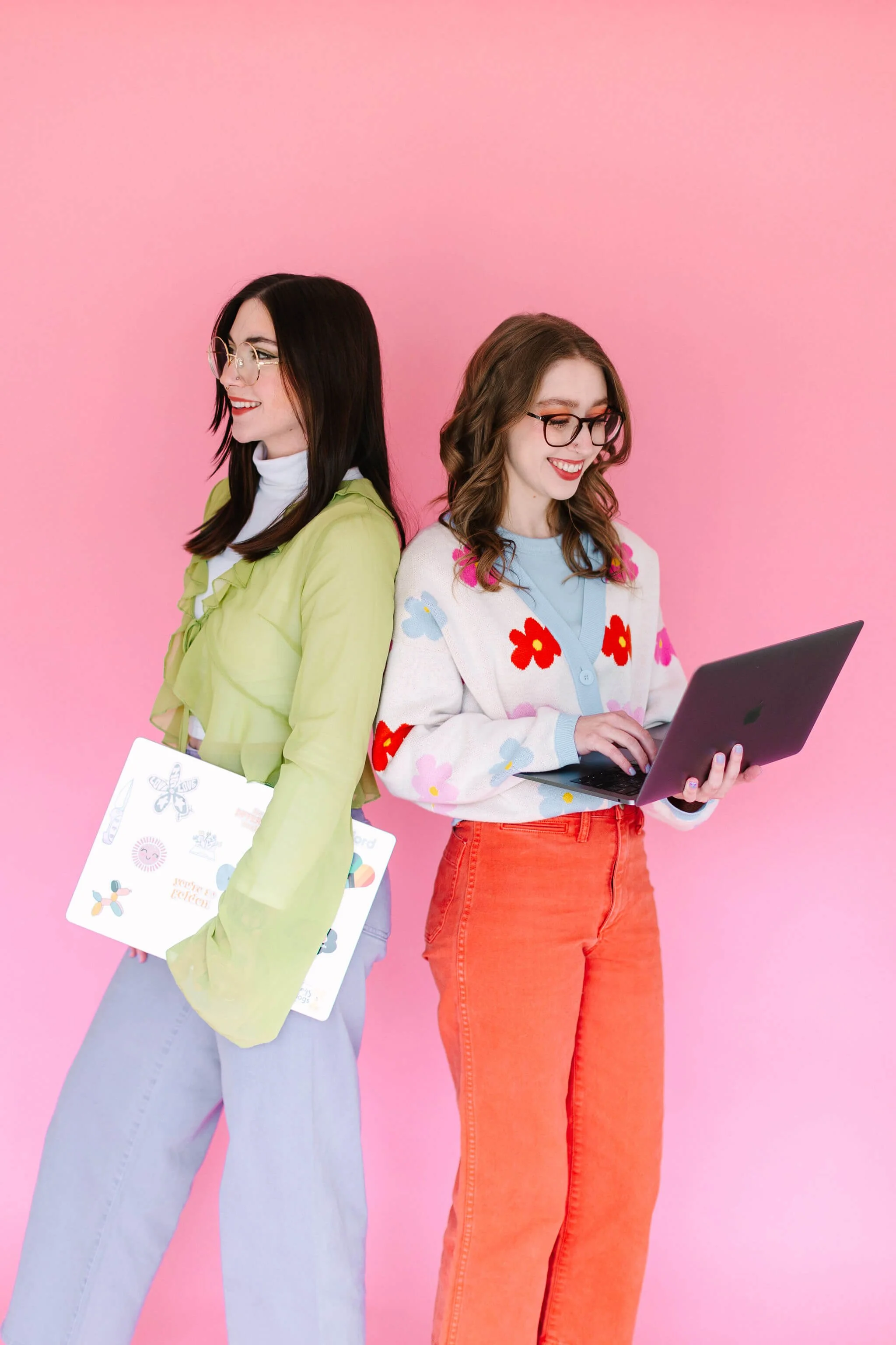 Logan and Emily stand in front of a pink background, holding their laptops.