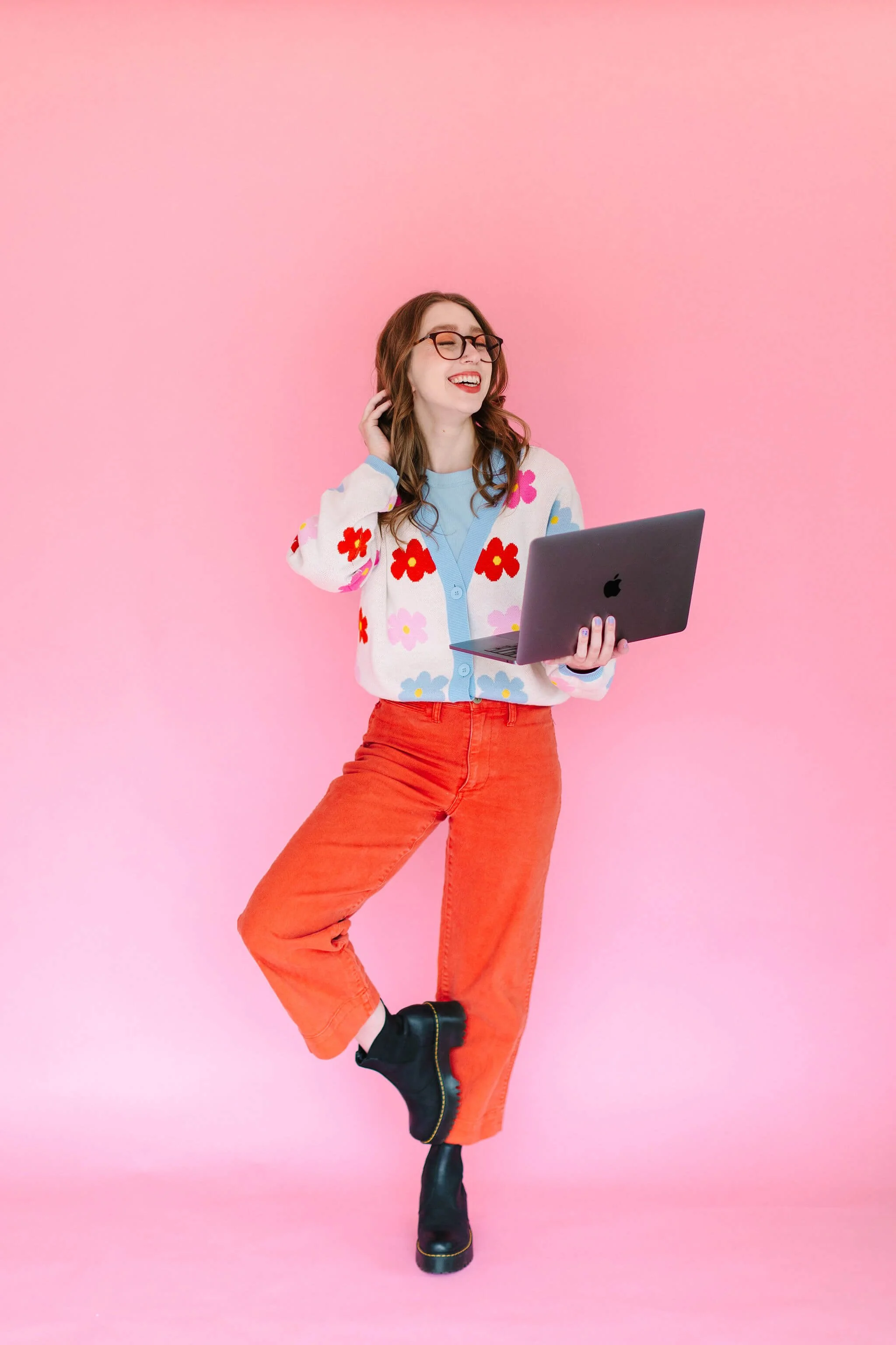 Logan stands in front of a light pink backdrop, holding her open laptop and smiling.