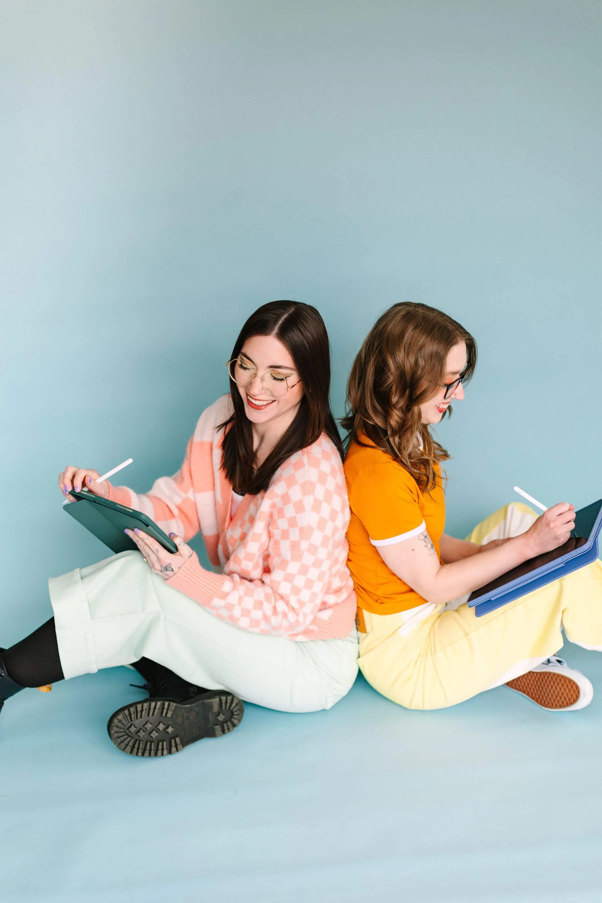 Logan and Emily sit on the floor back-to-back, writing on iPads and smiling.