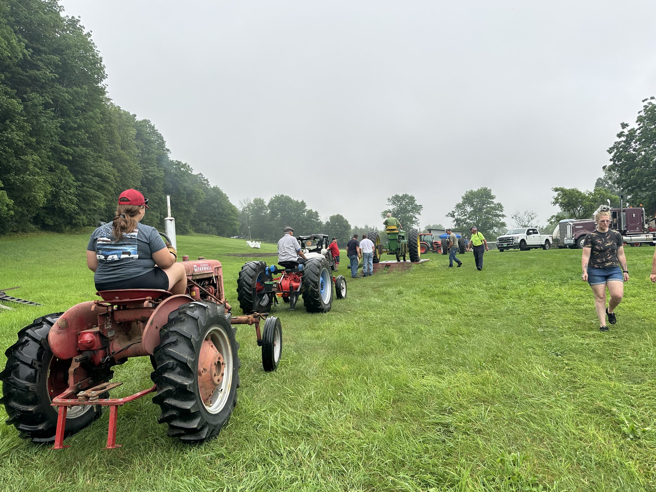 People gathered on a grassy field with vintage tractors on display, some walking and others sitting on tractors, under an overcast sky.