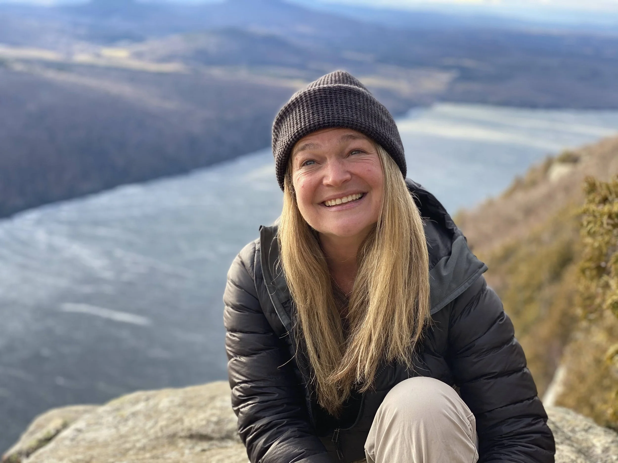 A woman smiling outdoors on a mountain. She is wearing a black jacket and a dark gray beanie, with a scenic lake and mountains in the background.