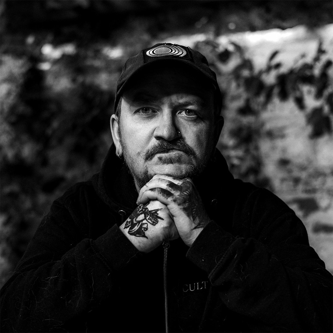 Schwarz-weiß-Porträt eines Mannes mit Tattoos, Bart und Kappe, der nachdenklich die Hände vor dem Kinn verschränkt, im Hintergrund eine Steilwand.