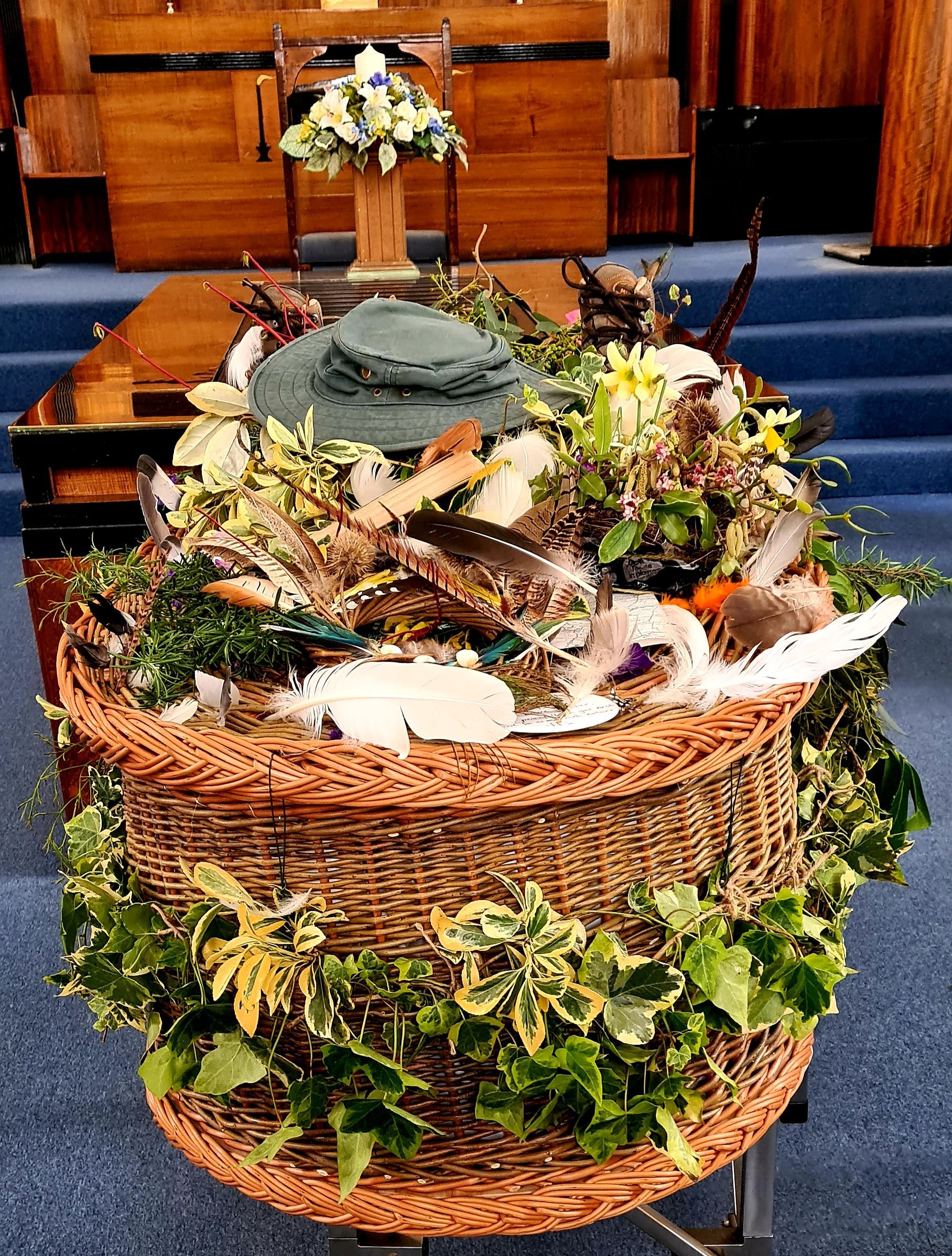 A wicker casket adorned with green foliage, feathers, and a green hat, situated on a platform in a wooden interior space.