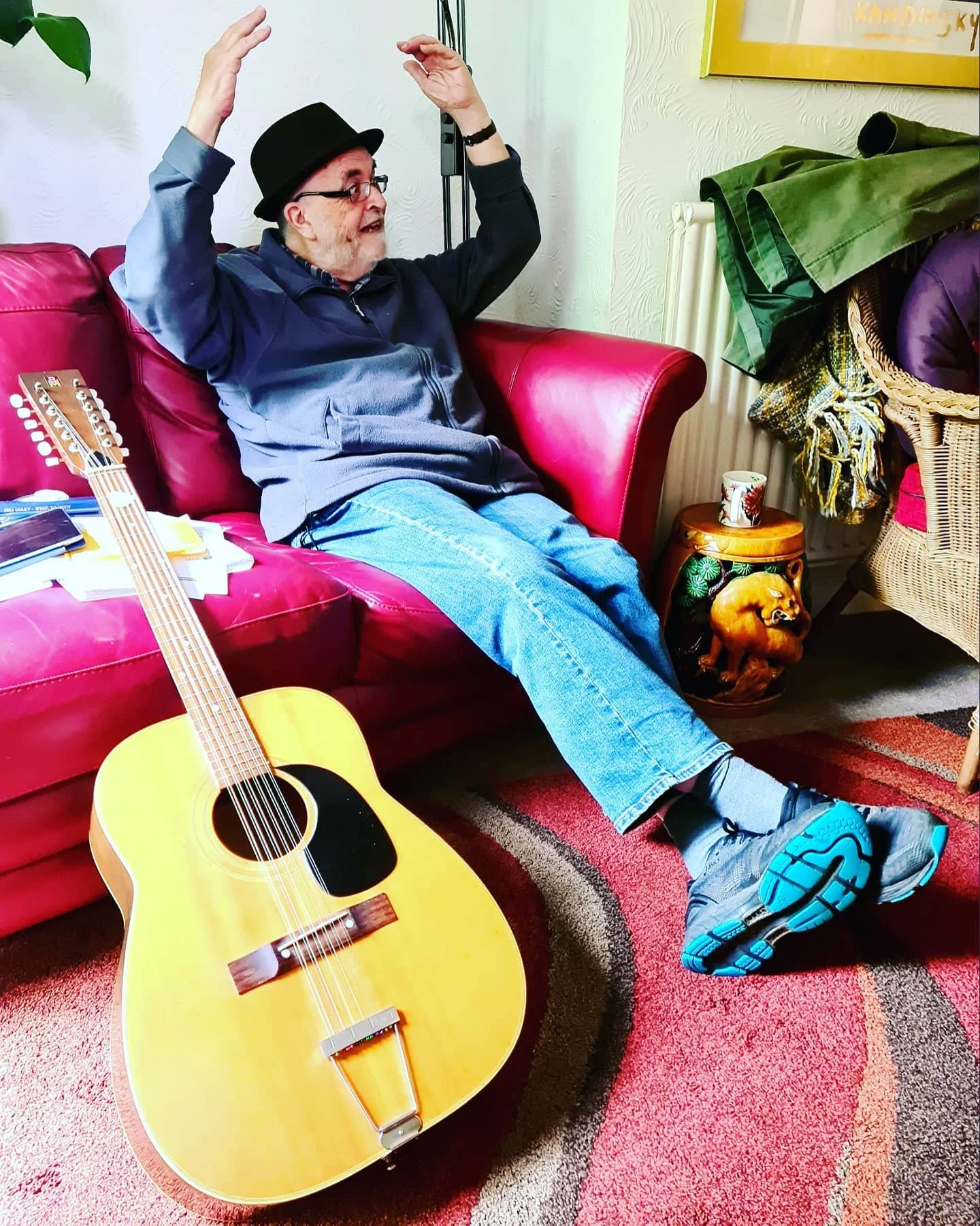 Man with black hat and glasses sitting on red couch, raising arms, next to acoustic guitar on floor.