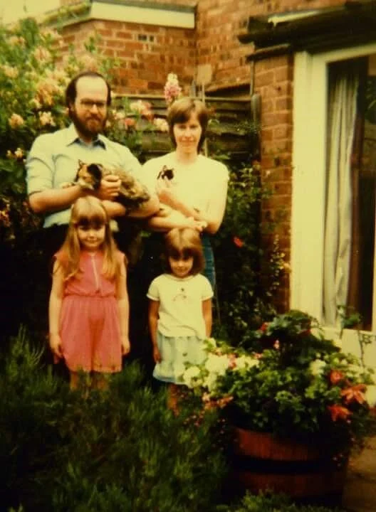 A family portrait in a garden, featuring an adult man and woman holding two cats, standing behind two young girls in dresses. The background includes brick walls, flowers, and greenery.