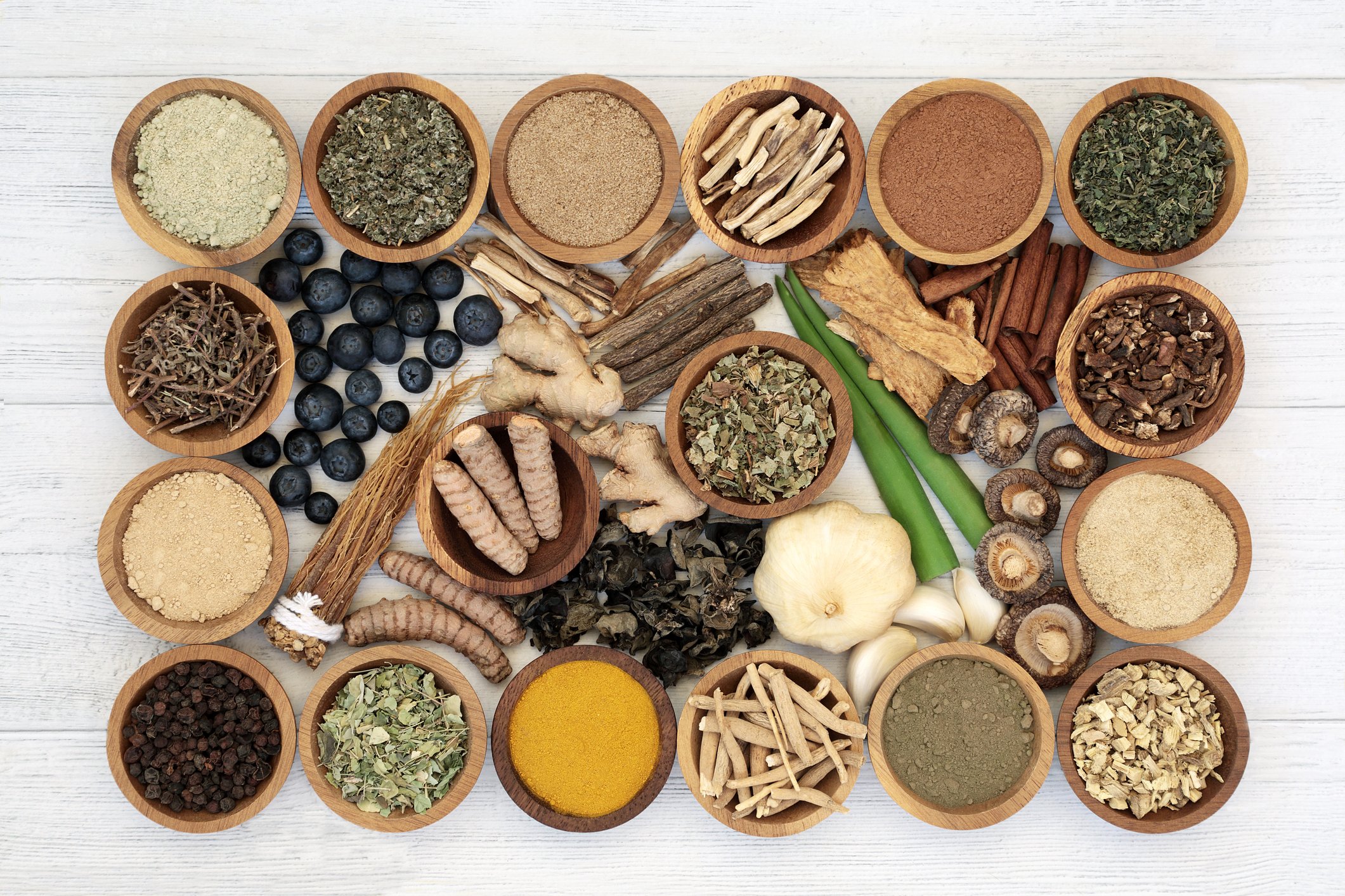 A top view of multiple small wooden bowls filled with various dried herbs, spices, and berries arranged on a white wooden surface.