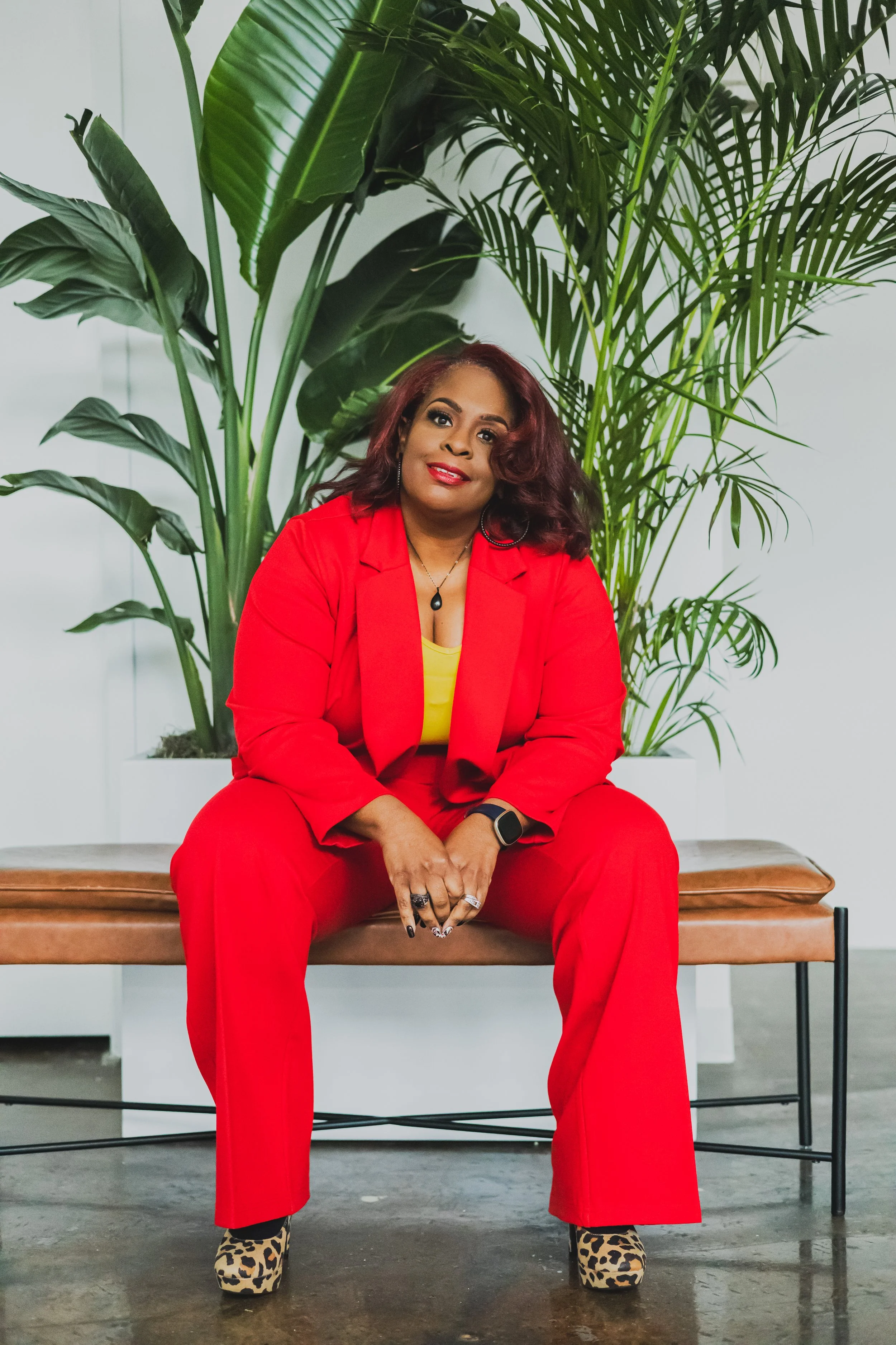 A woman with wavy, shoulder-length reddish hair, wearing a bright red suit with a yellow top underneath, sitting on a brown bench in front of large green plants in an indoor setting.