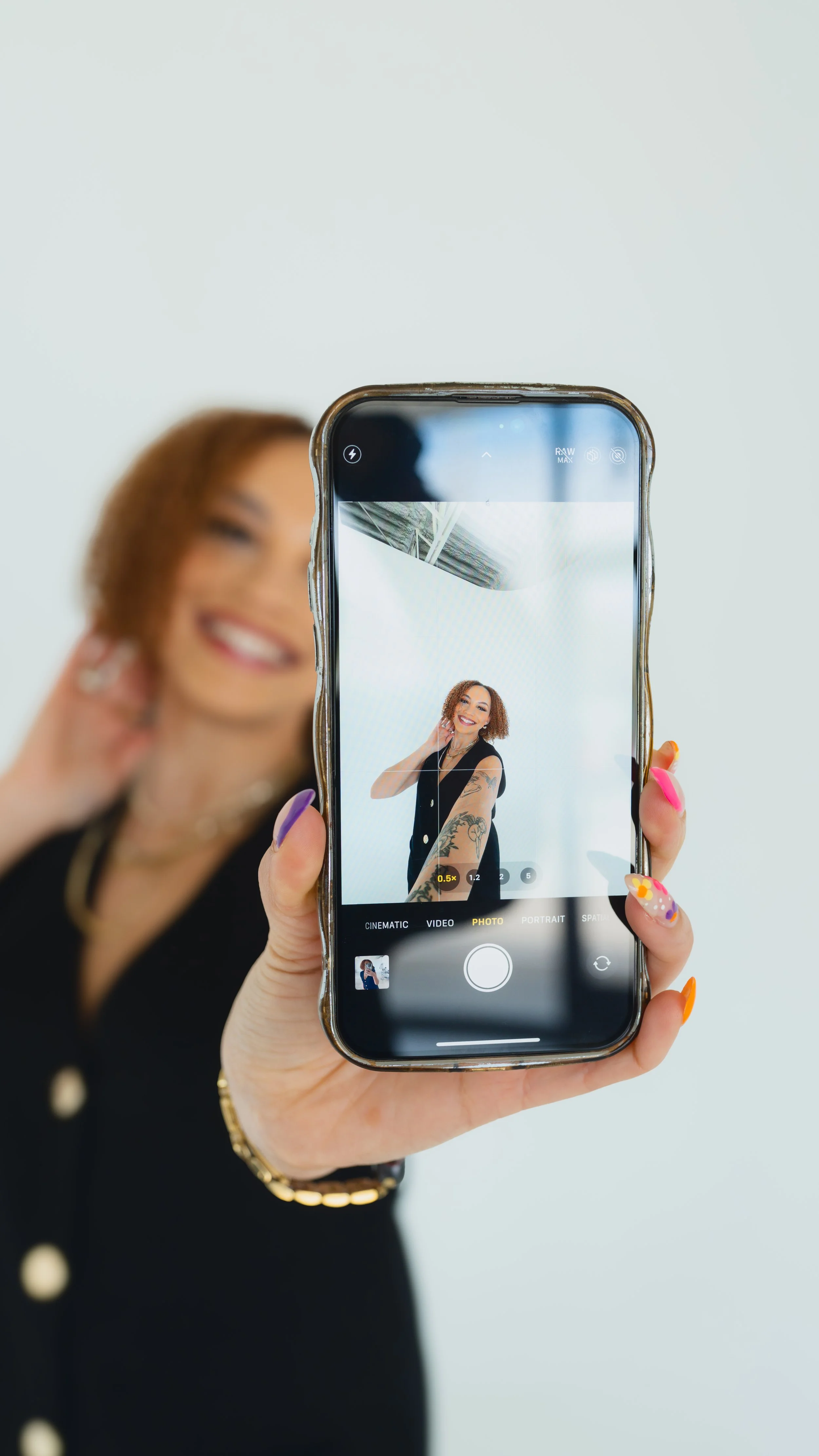 A woman with short curly hair smiling while taking a selfie with her smartphone, displaying her tattoos on her arm and wearing a black sleeveless top.