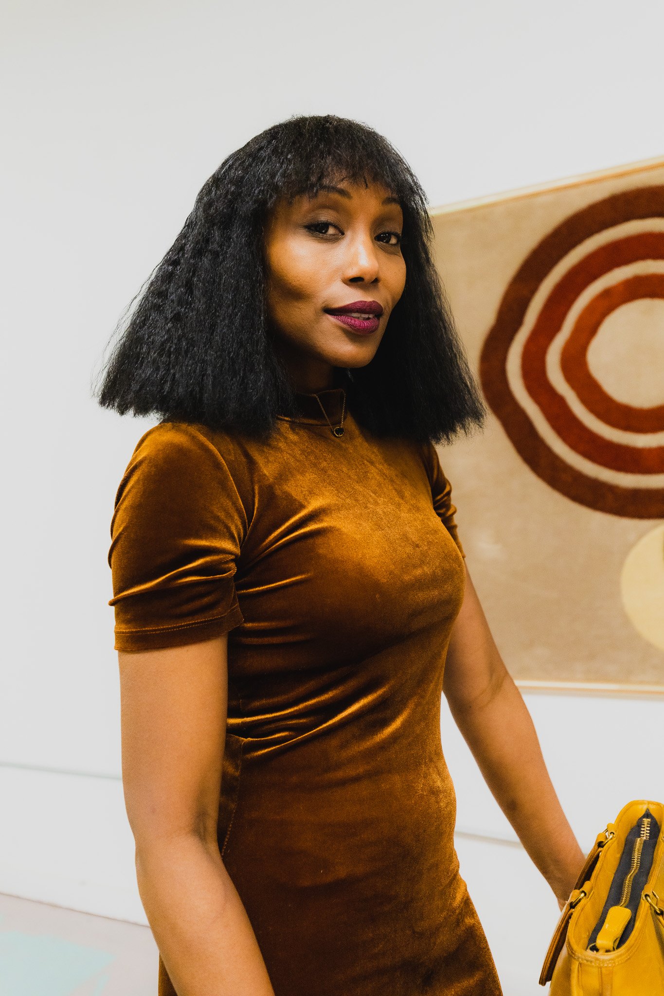 A woman with black, shoulder-length hair wearing a shiny, brown velvet dress, standing indoors in front of a white wall with a trashcan and a framed artwork in the background.