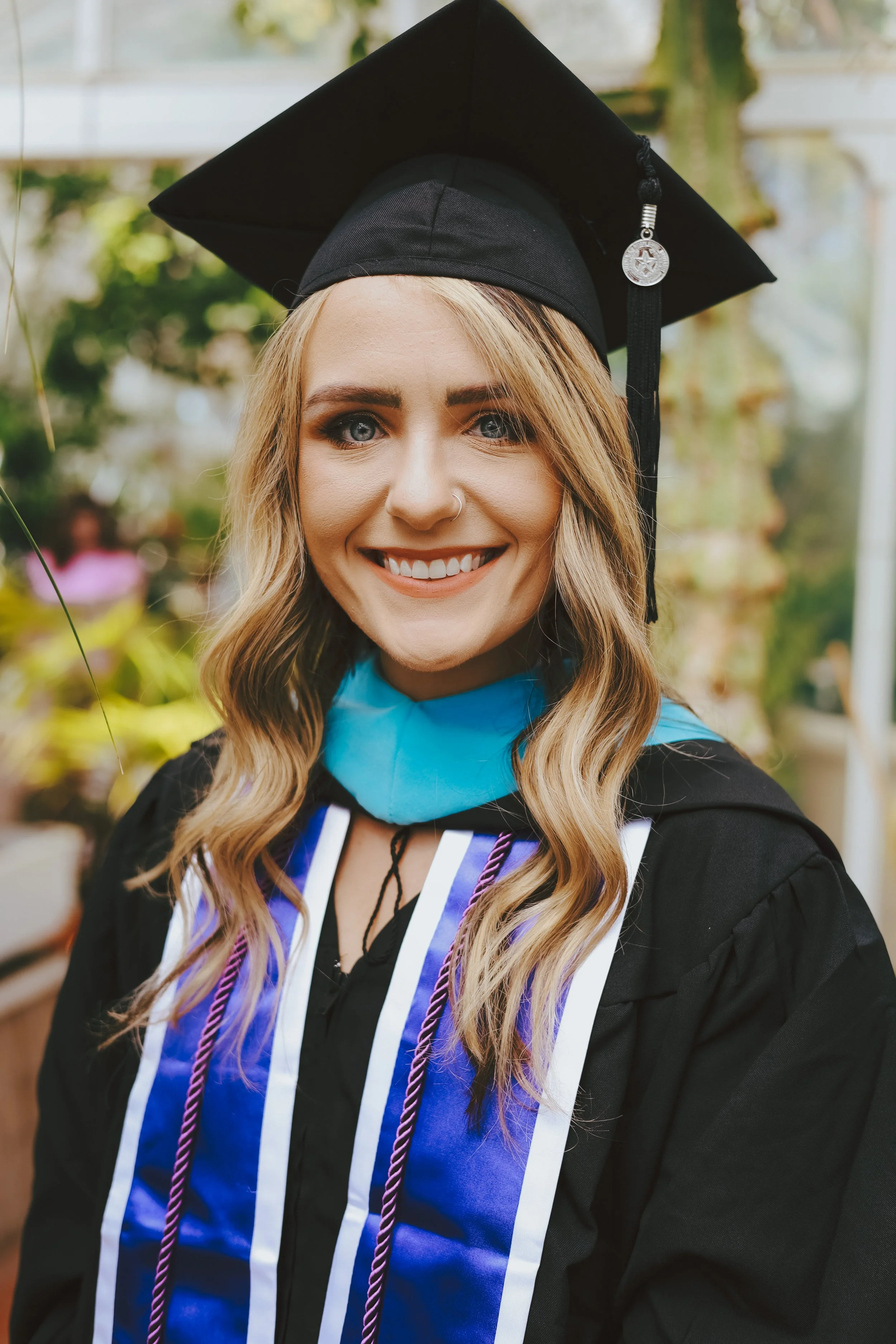 Smiling young woman in graduation cap and gown, outdoor setting with blurred greenery in the background.