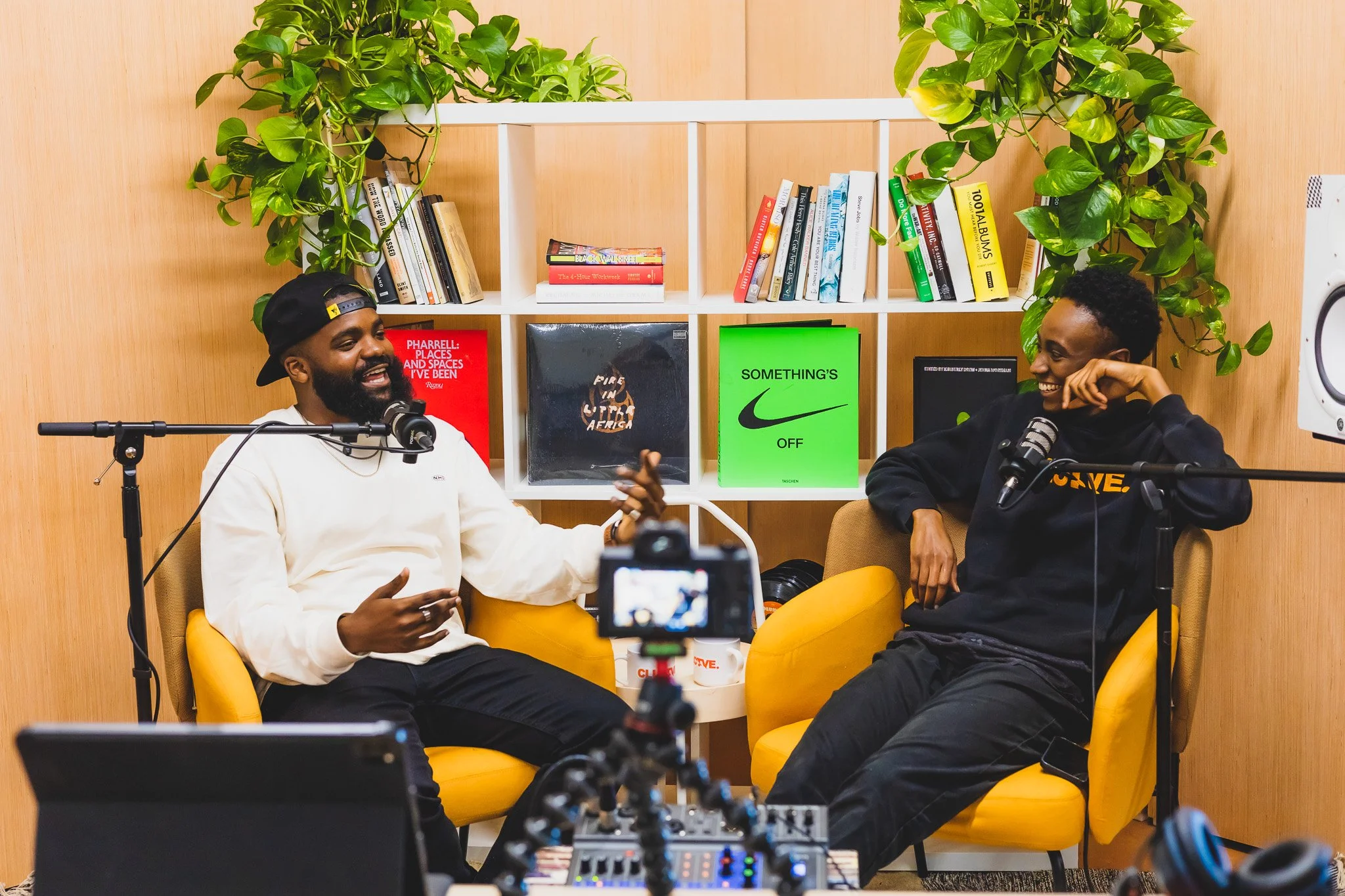 Two men are sitting in yellow chairs, laughing and talking during a podcast recording. The man on the left is wearing a white sweater and a black baseball cap backward, gesturing with his hands. The man on the right is wearing a black hoodie and glas
