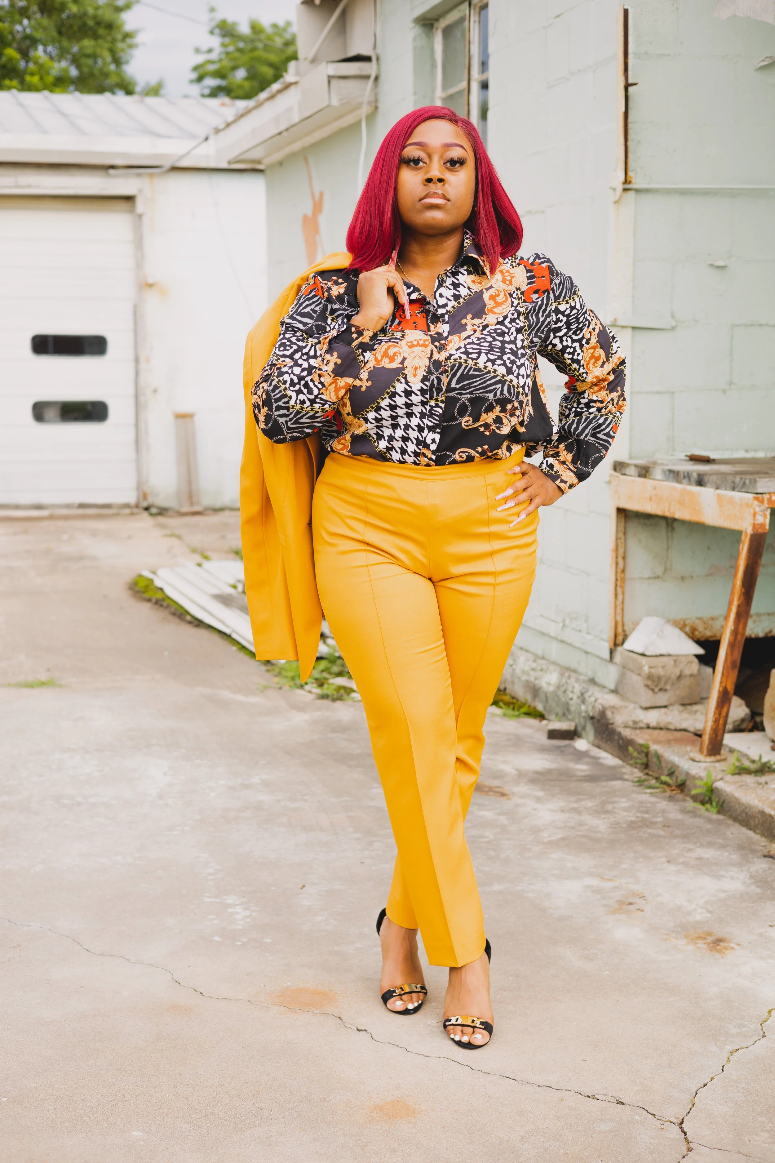 A woman with red hair wearing a colorful patterned blouse and yellow pants, standing outdoors on a cracked concrete surface, holding a yellow jacket over her shoulder.