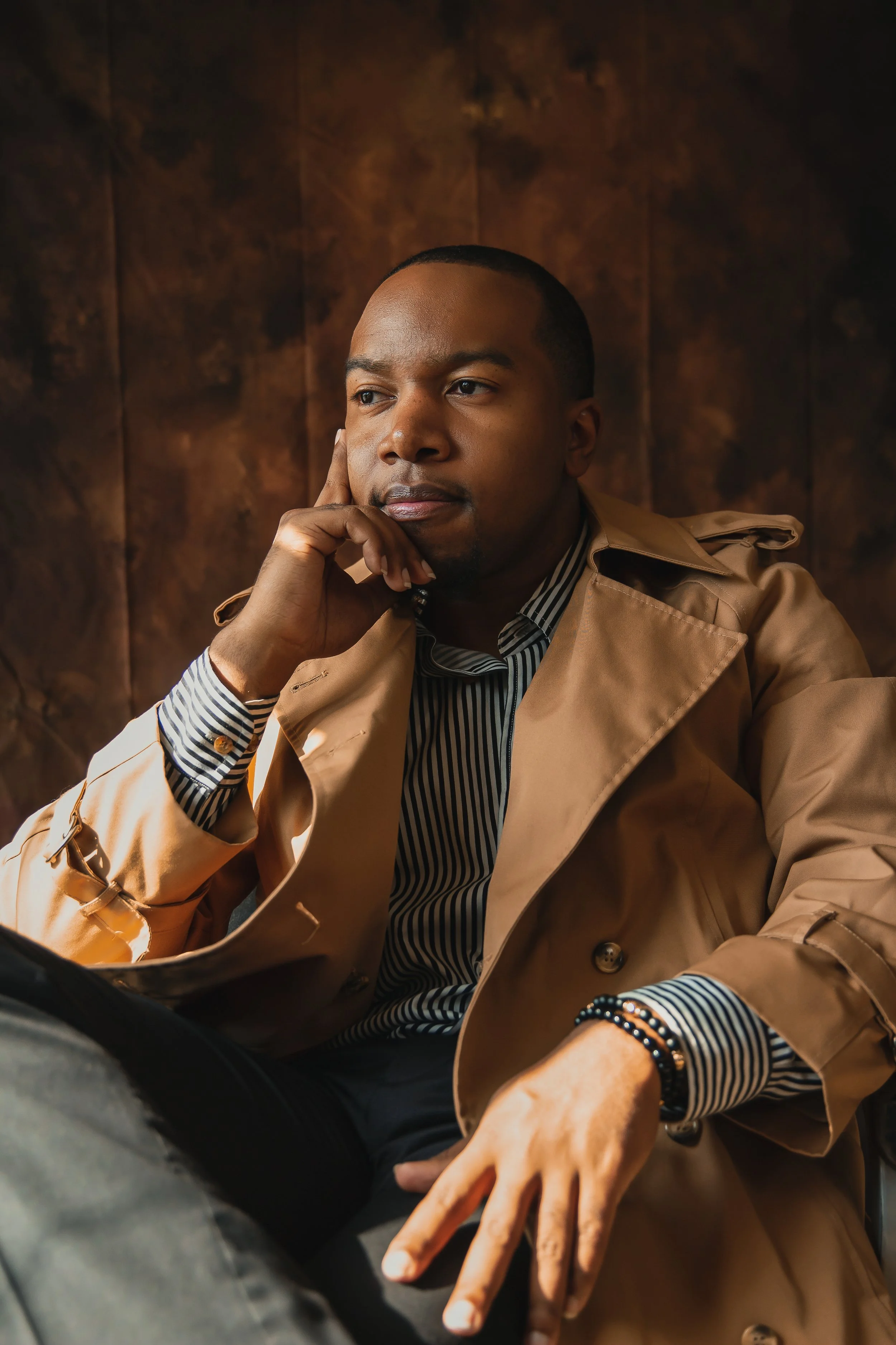 A young man in a beige trench coat and a striped shirt, sitting with his hand resting on his face, captured in a contemplative pose against a dark wood-paneled background.