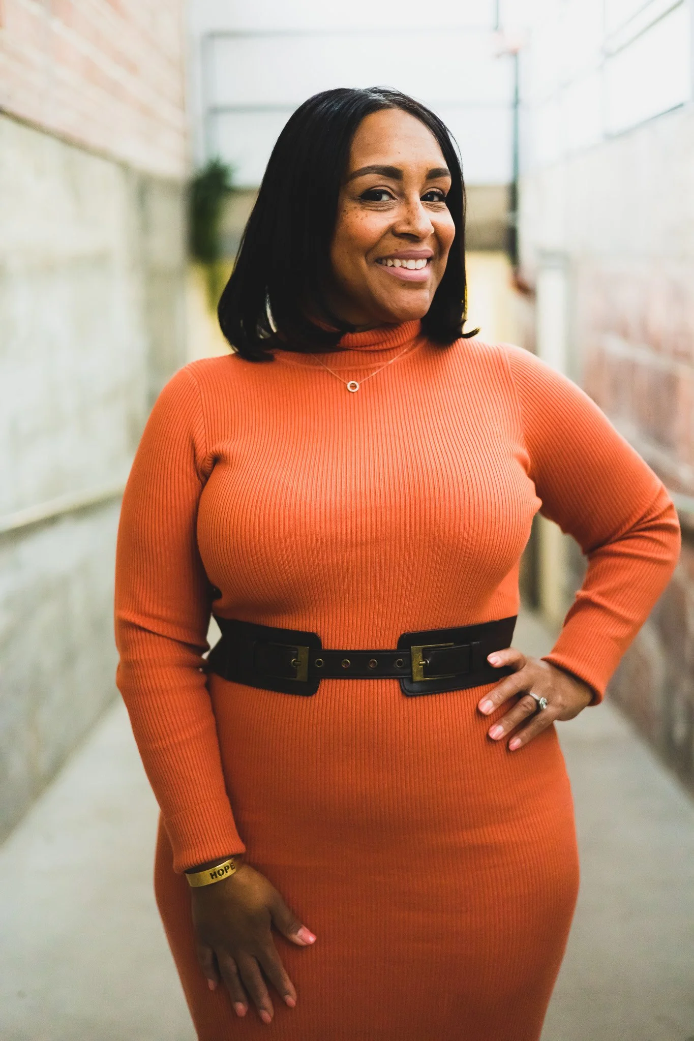 A woman with medium-dark skin, black hair, and a friendly smile, wearing an orange ribbed turtleneck dress with a black belt, standing in an industrial or subway station corridor.