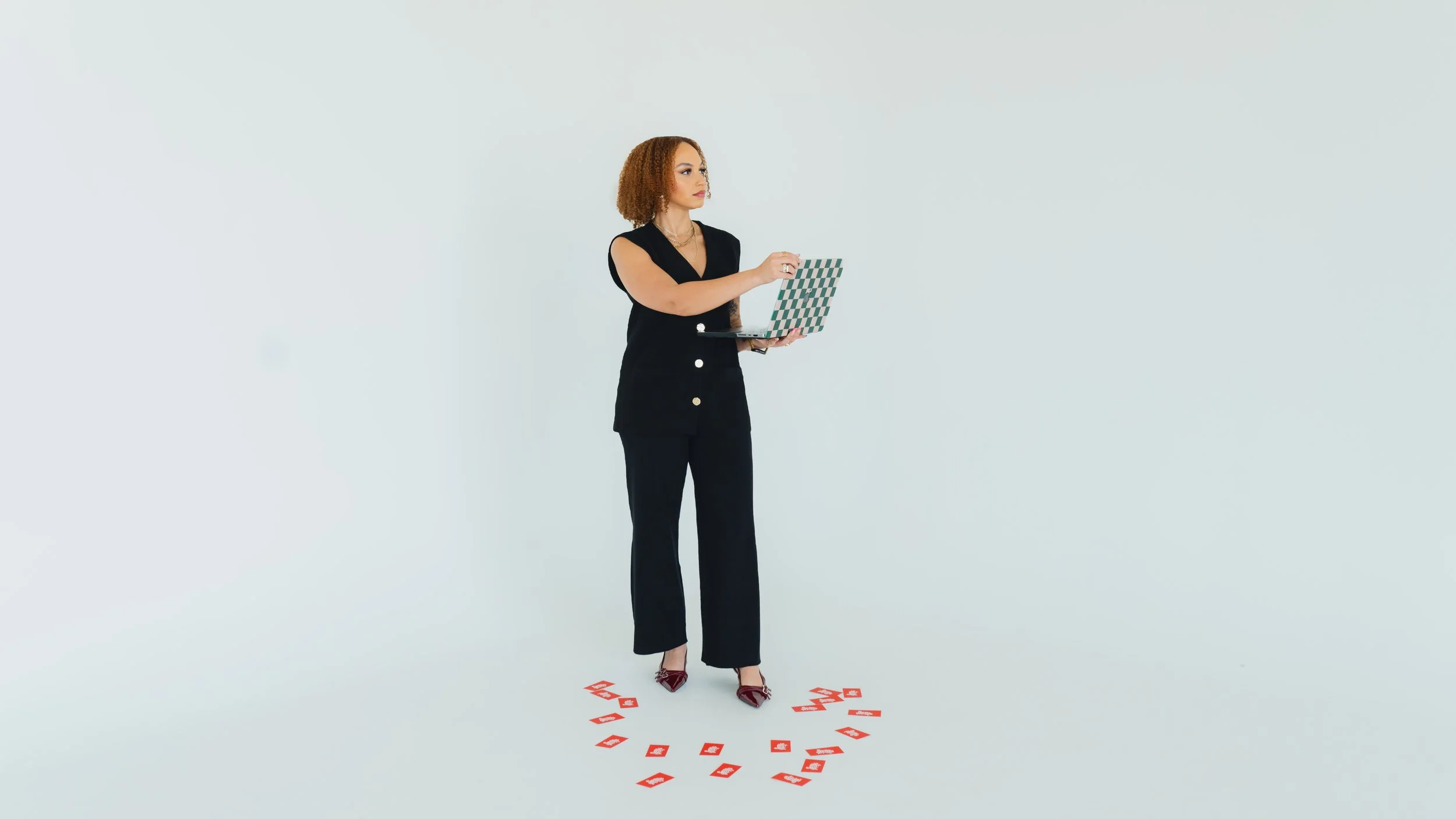 A woman with curly red hair wearing a black outfit holding a laptop and standing on a white background. There are red cards arranged in a circle on the floor.
