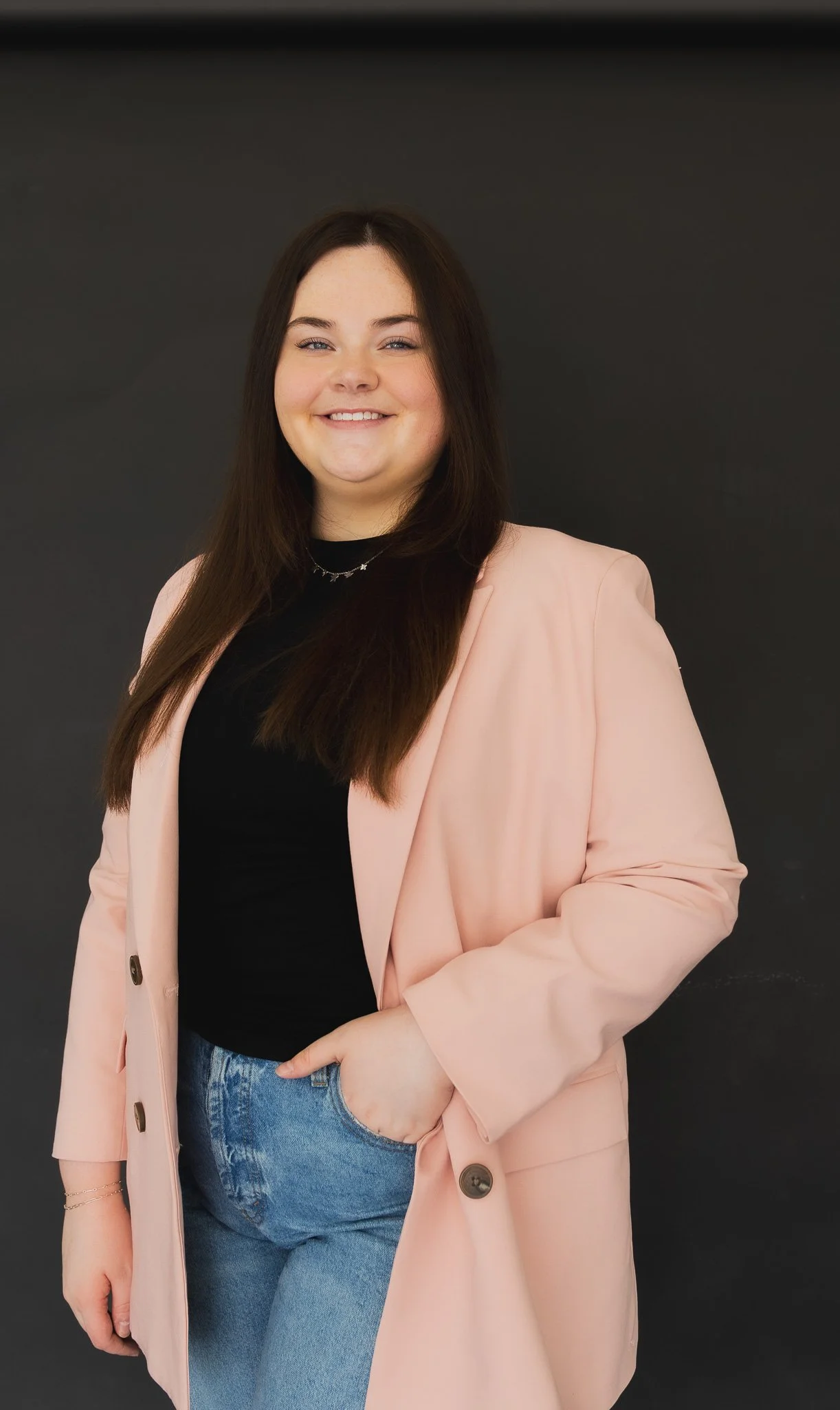 A woman with long dark brown hair, wearing a light pink blazer over a black top, and blue jeans, standing against a dark background. She is smiling with her left hand in her pocket.
