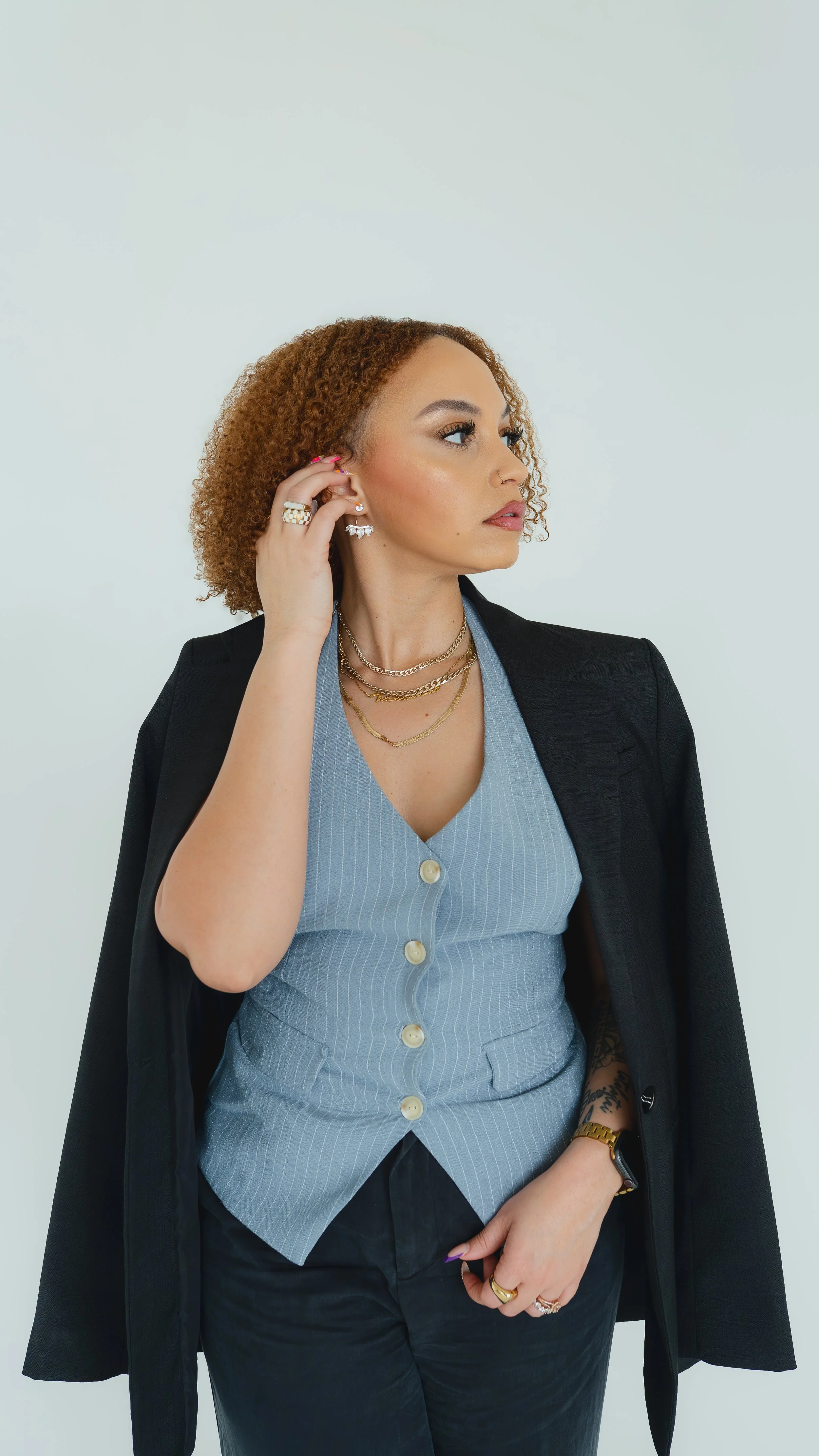 A woman with curly hair wearing a black blazer over a light blue button-up shirt, accessorized with layered necklaces, multiple rings, and earrings, posed against a plain light background.