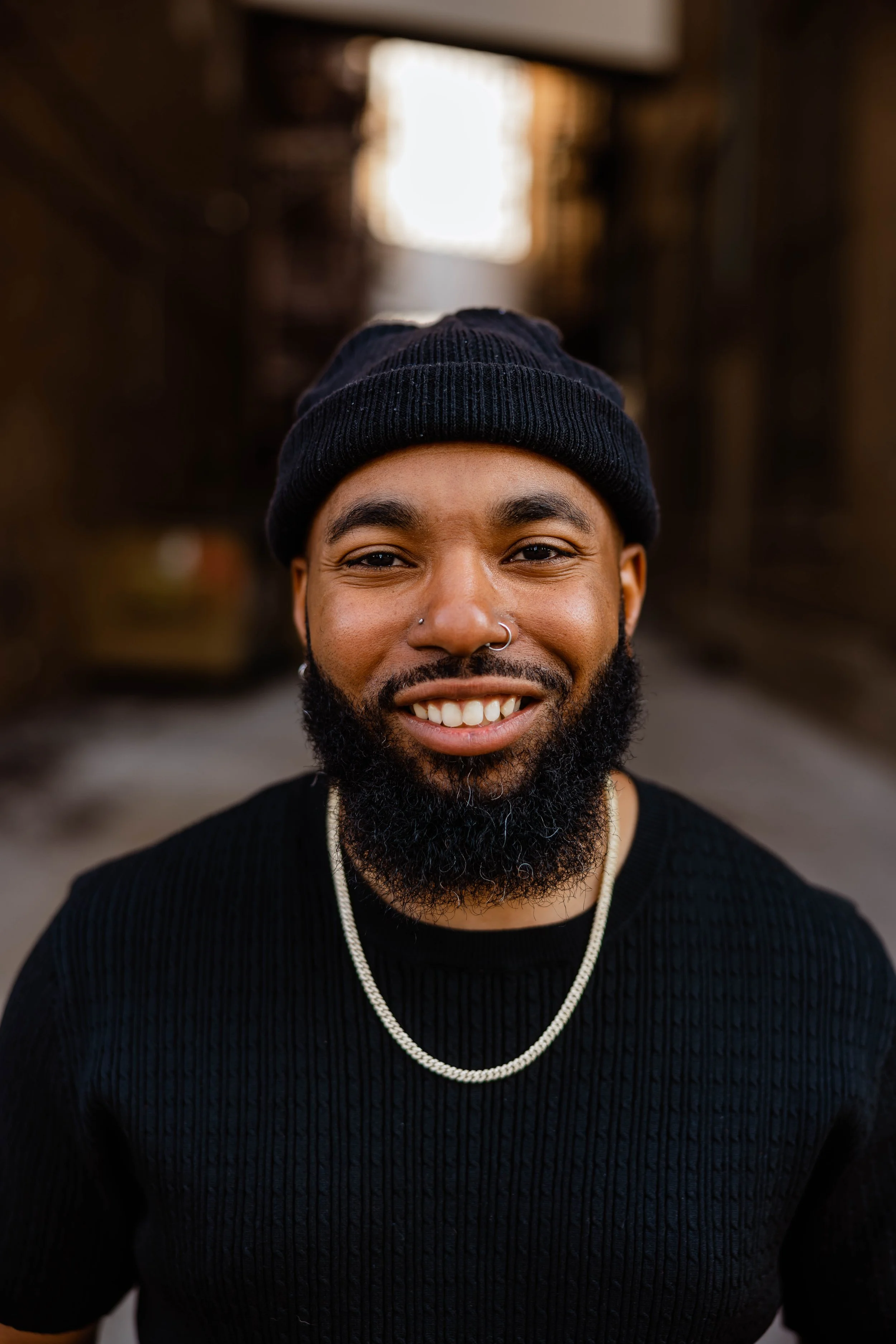 Close-up of a smiling man wearing a black beanie and a black sweater, with a silver chain necklace, outdoors with blurred background.